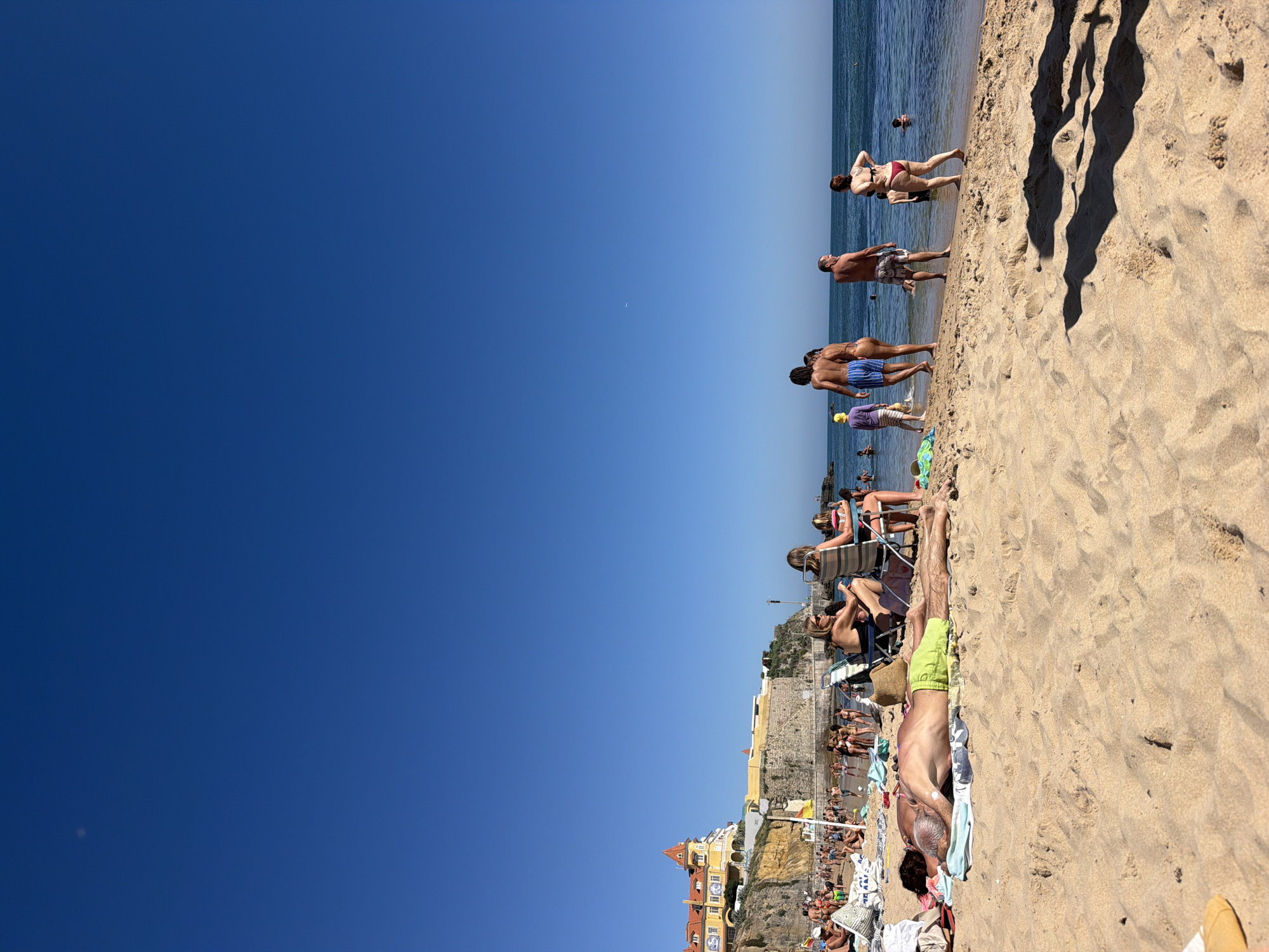 The image depicts a crowded beach scene in Cascais, Portugal. The sun is shining brightly, indicating it's likely daytime, and the weather appears to be warm and clear.
The foreground is dominated by a sandy beach with numerous people sunbathing and relaxing. Several individuals are lying on towels, with one man in the foreground positioned supine, wearing green shorts. Further up the beach, groups of people are standing and walking, some appearing to be in or near the water, which is calm and blue. The water's edge is lined with many beachgoers.
In the middle ground, a row of beach chairs is set up, with some people seated and others standing. Behind the beachgoers, a cliff face rises, topped by buildings and structures, including yellow and orange buildings with red-tiled roofs. These buildings suggest an urban or town setting adjacent to the beach. The architecture hints at a European coastal location.
In the background, the ocean stretches to the horizon under a clear blue sky. The shadows cast on the sand indicate the sun is positioned high, likely mid-day. The overall mood appears relaxed and summery, with people enjoying a day at the beach. There is no visible text in the image.