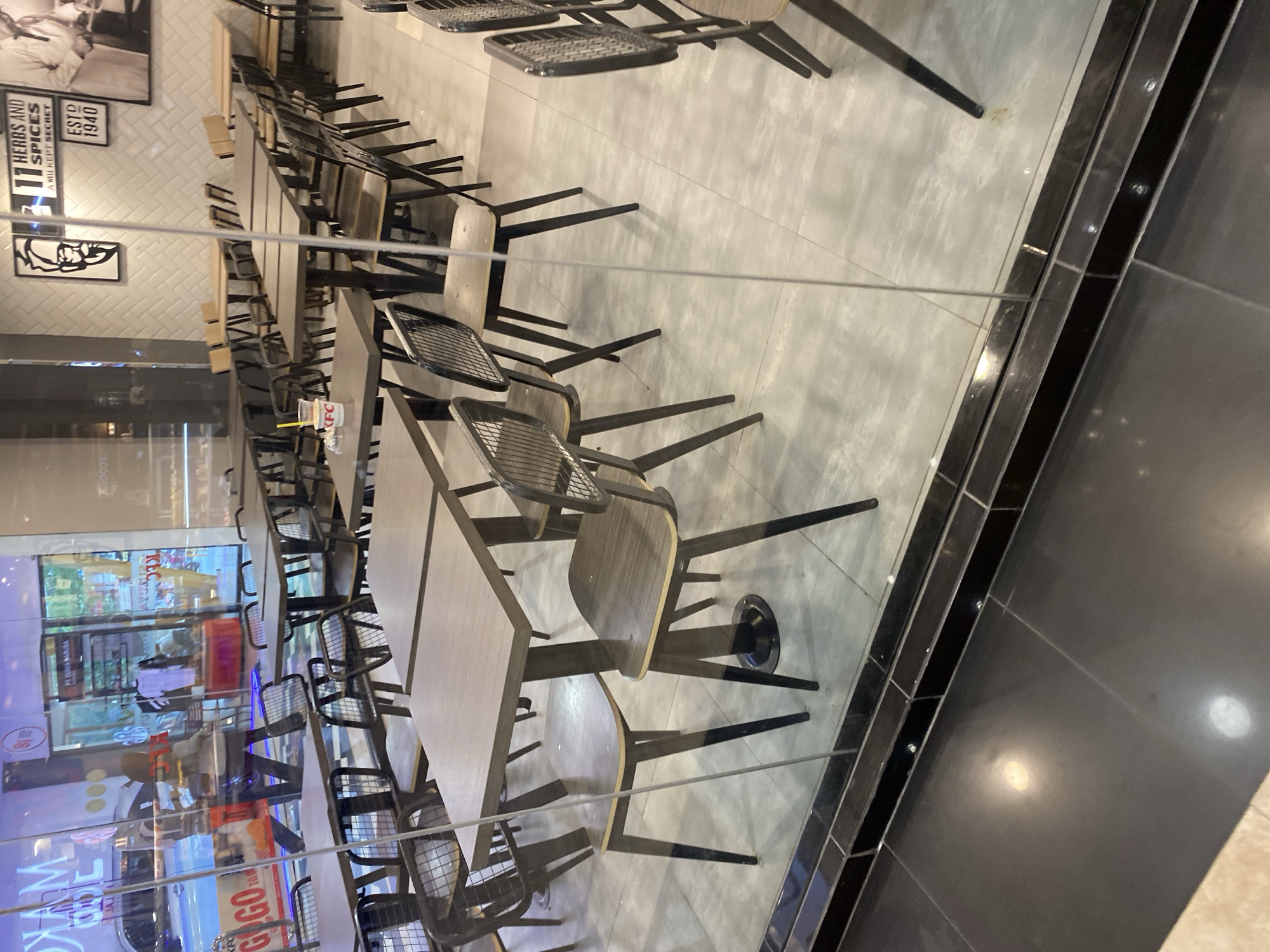 The image shows the interior of a KFC restaurant in Banda Aceh, Indonesia, viewed through a glass storefront. The restaurant is empty of customers. Several tables and chairs are visible, arranged in rows. Some tables are empty, while others have a cup and straw on them. The chairs are made of metal and wood. The walls are decorated with KFC branding, including a portrait of Colonel Sanders and the slogan "11 Herbs and Spices." The floor is tiled. Reflections of the street and people outside are visible in the glass.