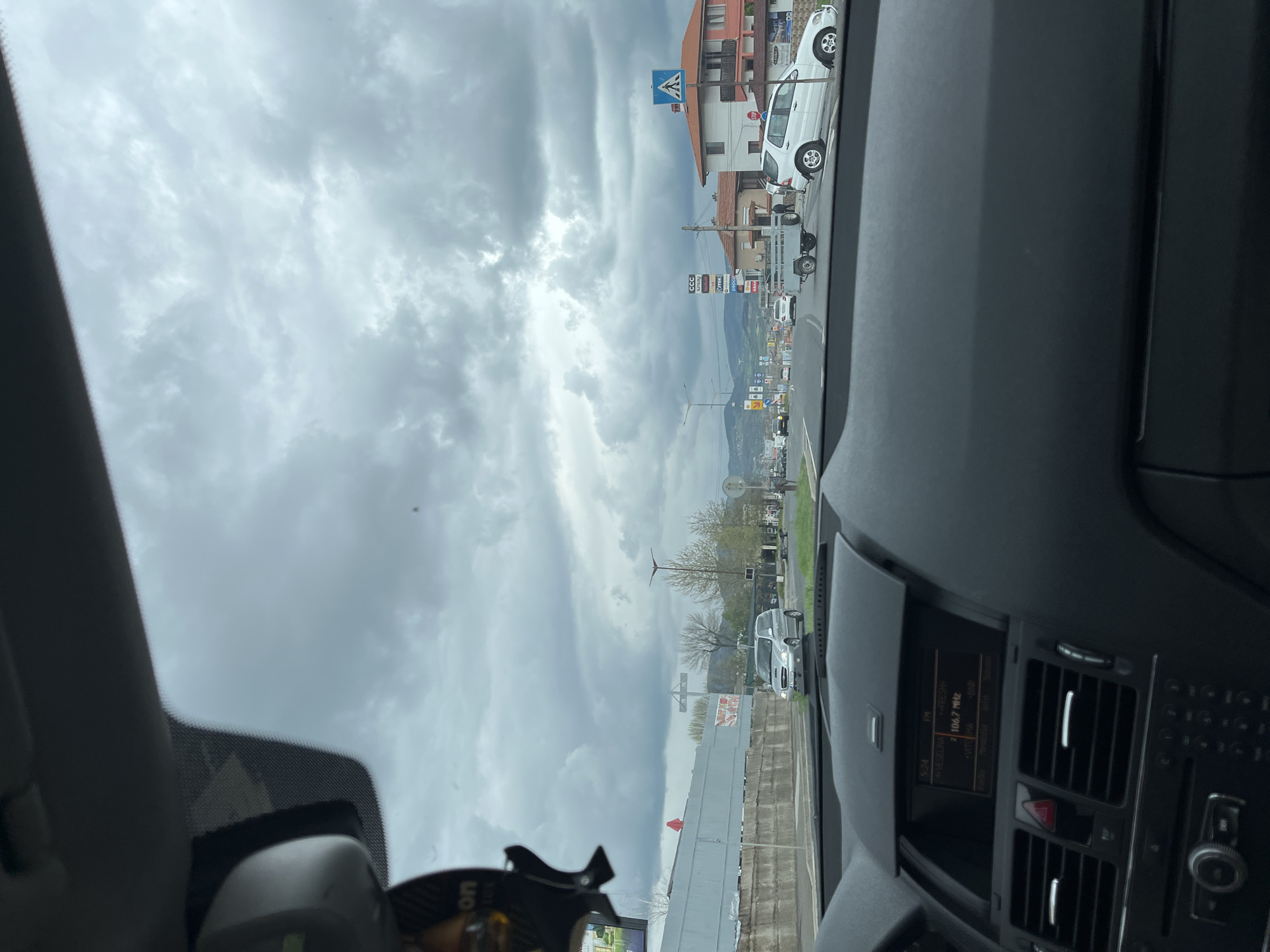The image is a landscape photograph taken from inside a car, looking out through the windshield onto a street scene under a heavily overcast sky.
In the foreground, parts of the car's interior are visible. On the right, a dark dashboard features air vents, a control panel with a red hazard light button, and a digital display showing "FM 106.7 MHz" and "Radio". To the upper left, a rearview mirror assembly and a car air freshener are partially visible.
The midground depicts a road with traffic moving or parked on the right side. Several buildings with red-tiled roofs and white facades line the street. A white SUV or van is parked near one of these buildings, and a small trailer is also visible. Various commercial signs are present, including "CCC Sprandi", "CORS", and "PEPC". A blue square road sign, indicating a pedestrian crossing, is prominently displayed.
In the background, distant hills or mountains are discernible beneath a vast, gray, and cloudy sky, suggesting a gloomy or potentially rainy day. Trees are scattered along the roadside in the distance. The scene captures an ordinary daytime moment from a vehicle, with no specific human activity highlighted.