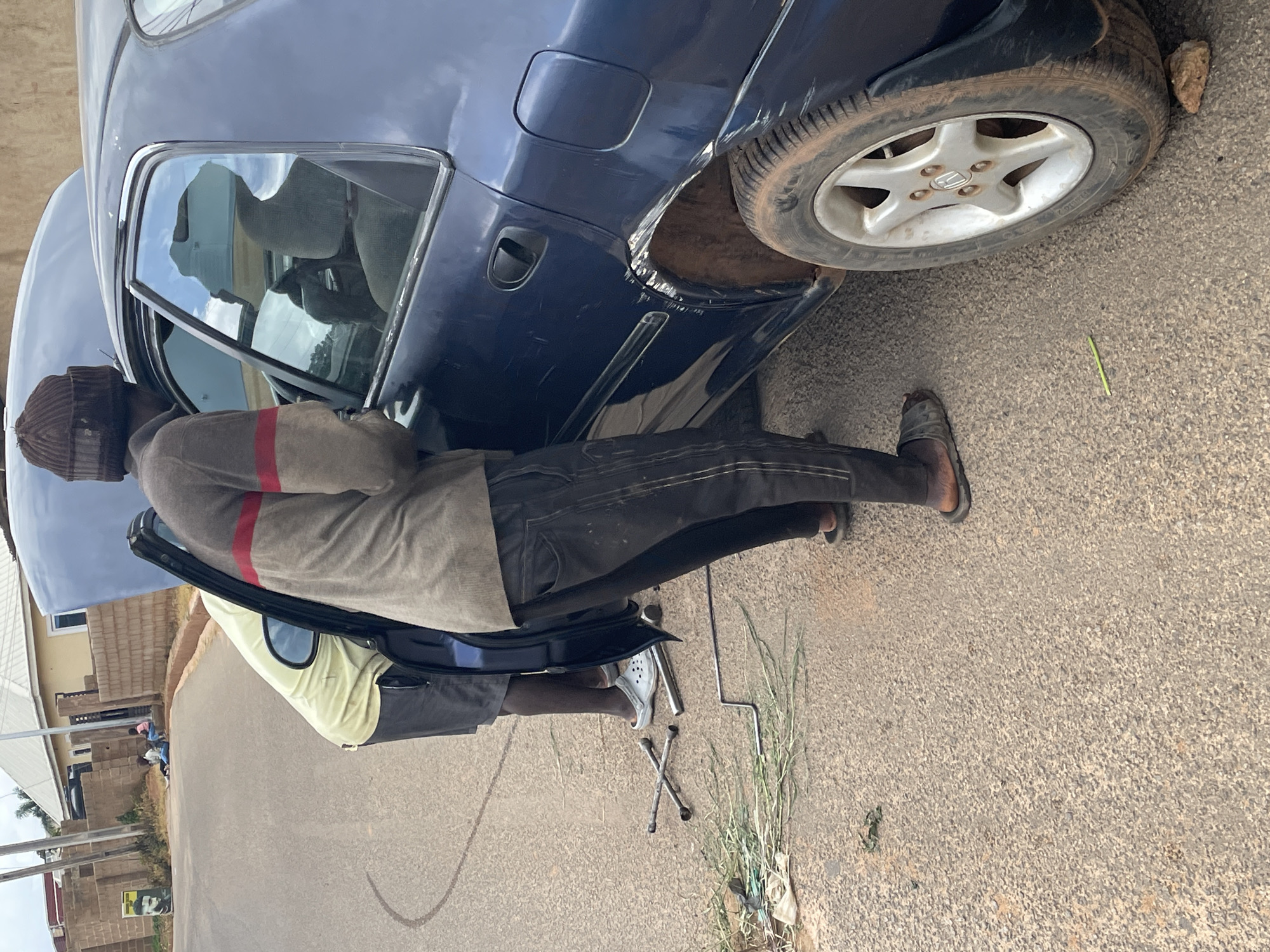 The image shows a person working on a dark blue car, which appears to be undergoing a tire change. The car is parked on a paved road, likely in Gura Top, Nigeria. The person is wearing a brown beanie, a gray sweater with red stripes, and dark pants. They are standing next to the open driver's side door, with their hands near the car's interior. A spare tire and a car jack are visible on the ground near the rear wheel, suggesting the car may have a flat tire. The sun appears to be shining, casting some shadows, indicating it is daytime. In the background, there are buildings, some of which appear to be residential, with a few other people visible in the distance. The scene is set outdoors, on what looks like a typical roadside in a populated area. The overall impression is of a practical, everyday event, likely a car repair being carried out by an individual.