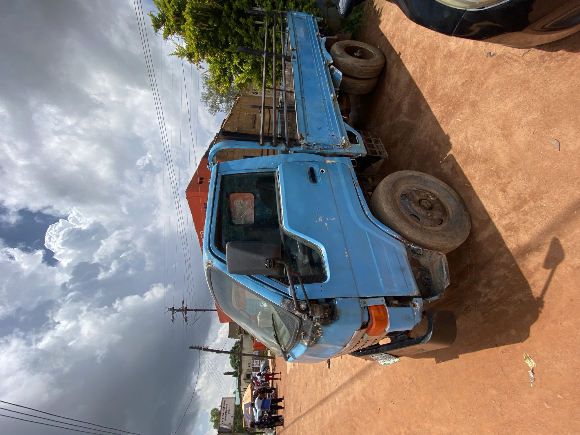 A bright blue flatbed truck is parked on a dirt road in Gura Top, Nigeria. The truck has a worn appearance, with visible rust and some damage to the front bumper and hood. The bed of the truck is empty, and it is supported by dual rear wheels on the right side. In the background, an orange building stands with a few trees and utility poles. The sky is filled with dramatic, heavy clouds. Several people are visible in the distance on the left side of the image, near a stall with umbrellas. The ground is a reddish-brown dirt.