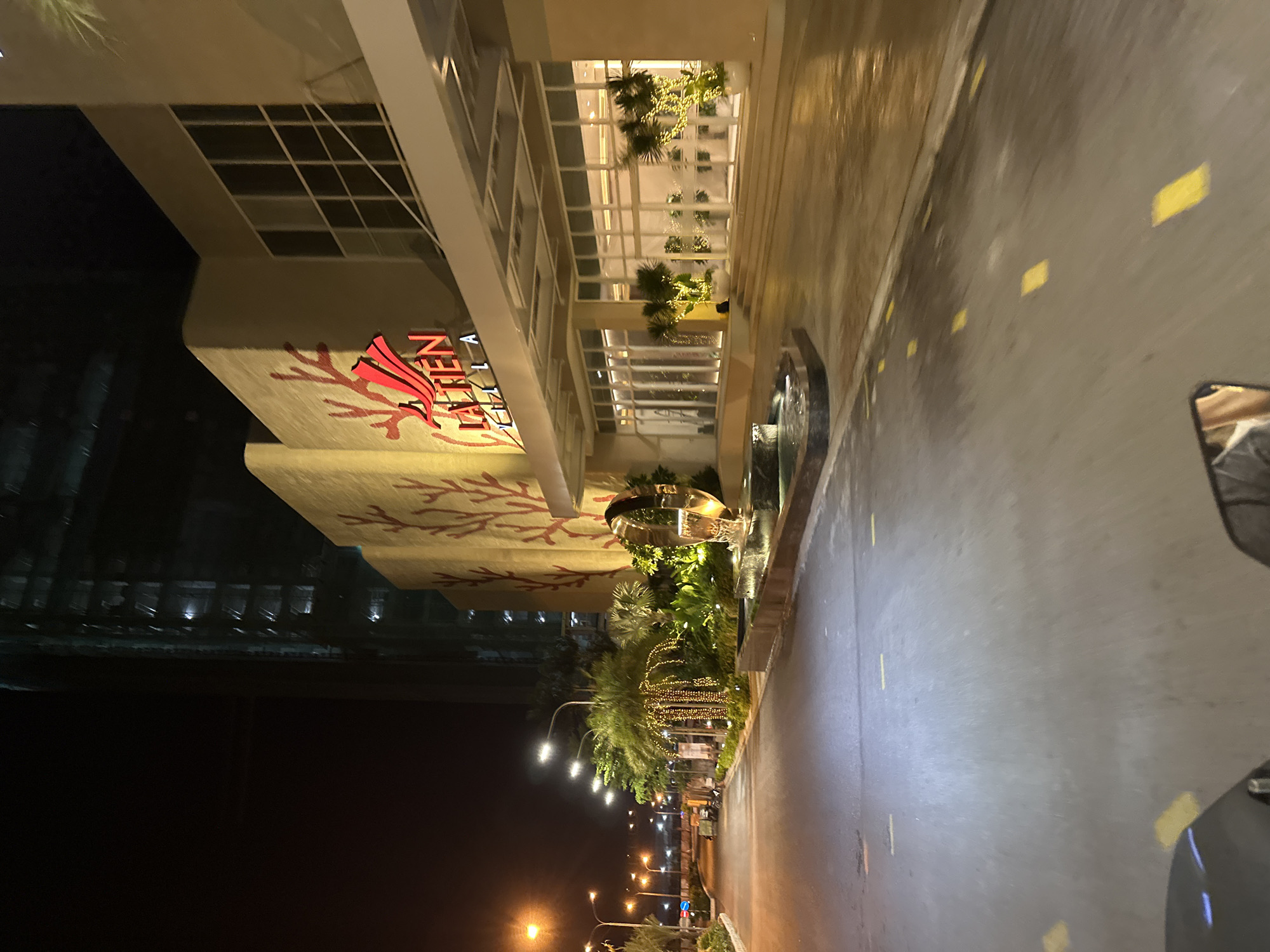 The image captures a nighttime scene in front of a modern building, possibly a hotel or residential complex, located in Nha Trang, Vietnam. The building features large windows and is adorned with a distinctive red, stylized tree or coral-like motif on its facade. The text "LA TEN" is prominently displayed in red, angular lettering on the building's exterior, with "CITY" visible below it.
The setting is an outdoor area with a wet asphalt road, suggesting recent rain or damp conditions. Yellow rectangular markers are intermittently placed along the road. The scene is illuminated by streetlights, building lights, and decorative string lights that are wrapped around palm trees and other greenery, creating a festive atmosphere. A modern metallic sculpture, possibly a water feature or decorative element, is visible near the entrance, surrounded by lush tropical plants.
The time of day is clearly night, with the darkness of the sky contrasted by the artificial lighting. The overall mood is serene and inviting, with the lighting highlighting the architectural features and landscaping. There are no people visible in the frame, and the focus is on the building's exterior and the immediate surroundings. The presence of tropical plants and the architecture suggest a warm climate. A portion of a vehicle, likely a car, is visible in the lower right corner of the frame, reflecting some of the ambient light.