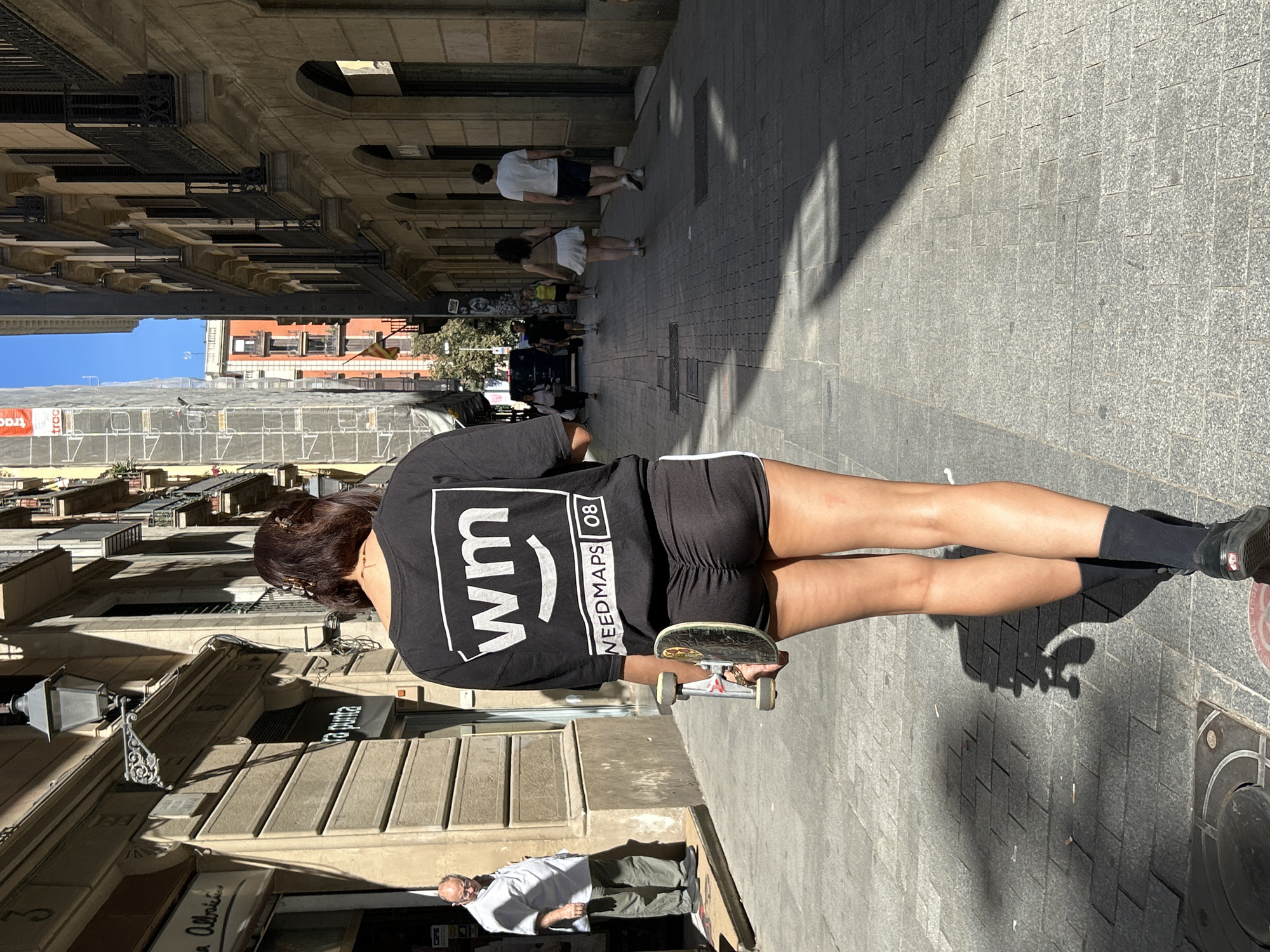 The image shows a young woman on a street in Barcelona, Spain, with a skateboard in hand. She is wearing a black t-shirt with the word "WEEDMAPS" and the number "08" on it, black shorts with white piping, black socks, and black sneakers. The street is paved with grey stones and lined with buildings. Other people are walking nearby, and a restaurant called “Tra Punta” is visible on the left of the shot. The lighting suggests it is a sunny day.