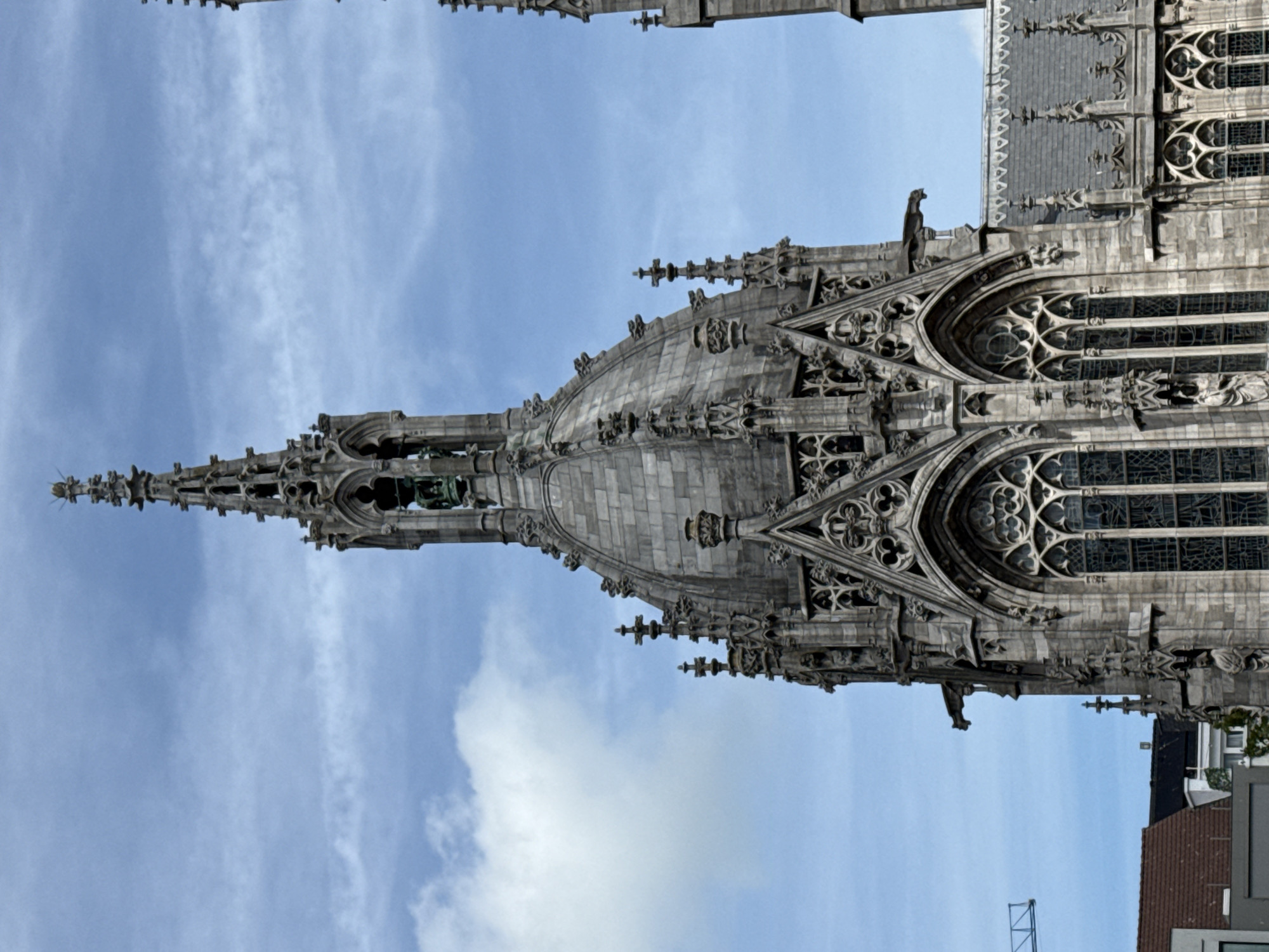 This is a close-up, low-angle, eye-level, outdoor, vertical shot of a gothic-style building spire against a cloudy blue sky. The spire is intricately detailed with stone carvings, small arched windows, and ornate finials. A bell is visible through an opening in the spire, and a gargoyle juts out from the side. To the right, another spire of a similar architectural style is partially visible. In the lower left of the frame, a crane is seen against the sky, and the corner of a tiled roof building is visible below. The lighting suggests it is daytime.