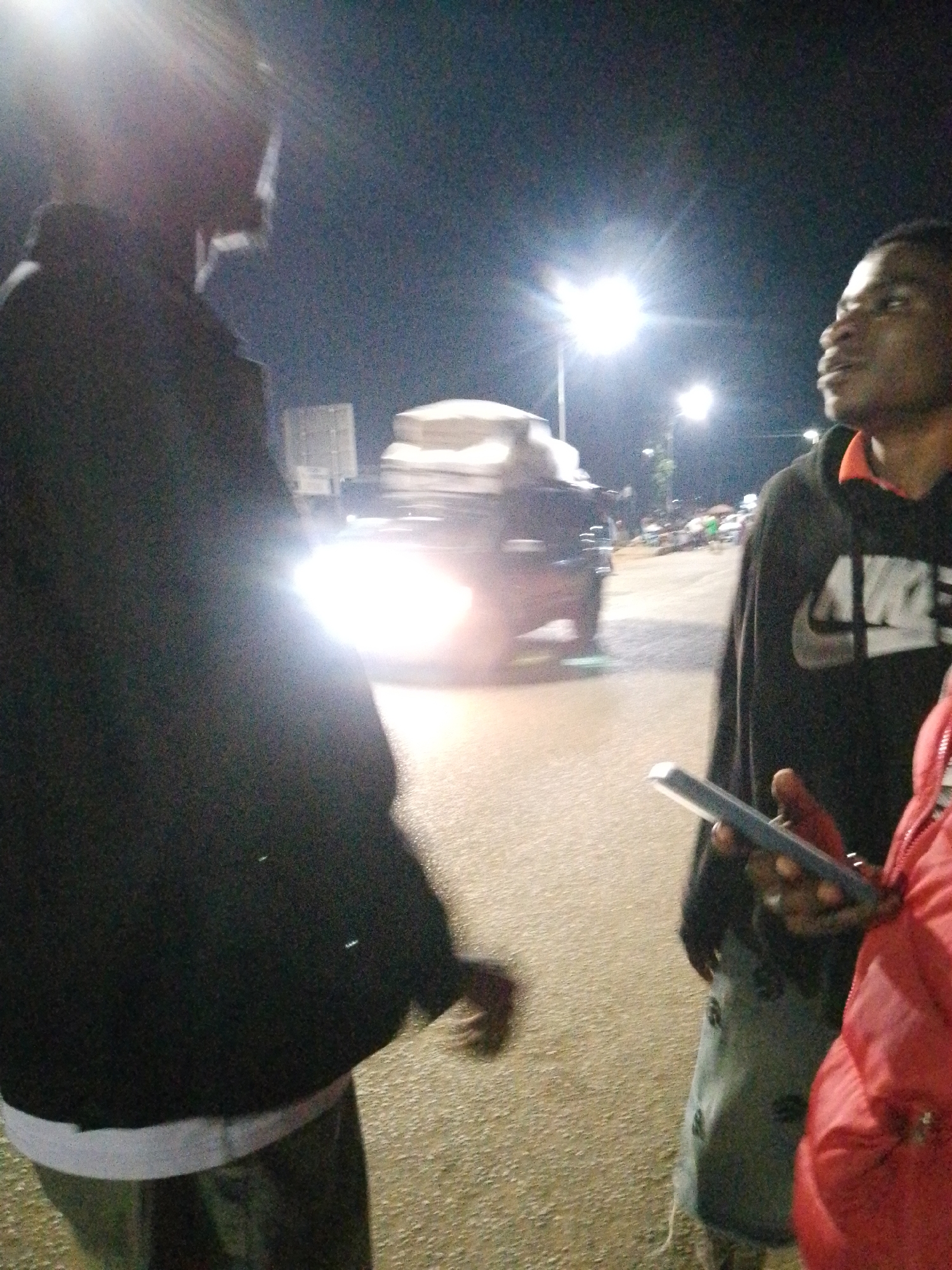 The image captures an outdoor scene at night in an urban environment. Two individuals are visible, with a third partially obscured. The foreground is dominated by the back of a person wearing a black hooded jacket over a white undershirt and dark pants. To the right, another person, wearing a black Nike hoodie and grey patterned pants, holds a smartphone. A red jacket is visible on a third person to the far right. In the background, a vehicle with illuminated headlights is in motion, and streetlights cast a bright glow, suggesting a roadway or parking area. The ground appears to be asphalt. The overall atmosphere is dimly lit, characteristic of nighttime urban lighting conditions.