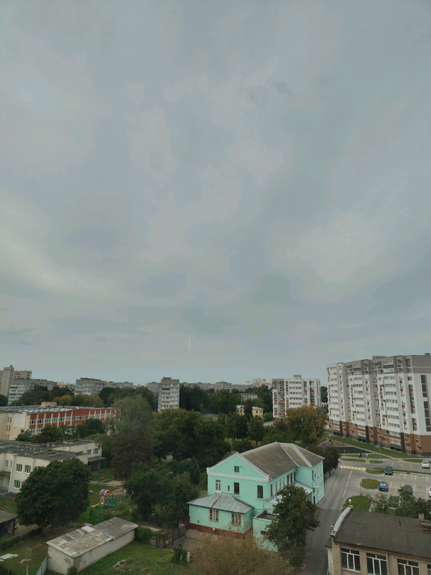 The image shows an aerial view of a residential area in Homyel, Belarus, under an overcast sky. In the foreground, a light blue building with a darker blue roof is visible, flanked by trees. To the left, a playground with colorful equipment is present. Beyond these, several apartment buildings of varying heights and architectural styles dominate the cityscape, many featuring balconies and light-colored facades. A road with parked cars and a pedestrian crossing is also visible, suggesting a typical urban environment. Trees and greenery are interspersed throughout the scene, indicating a mix of development and natural elements. The overall impression is of a moderately built-up urban area, likely a residential district.