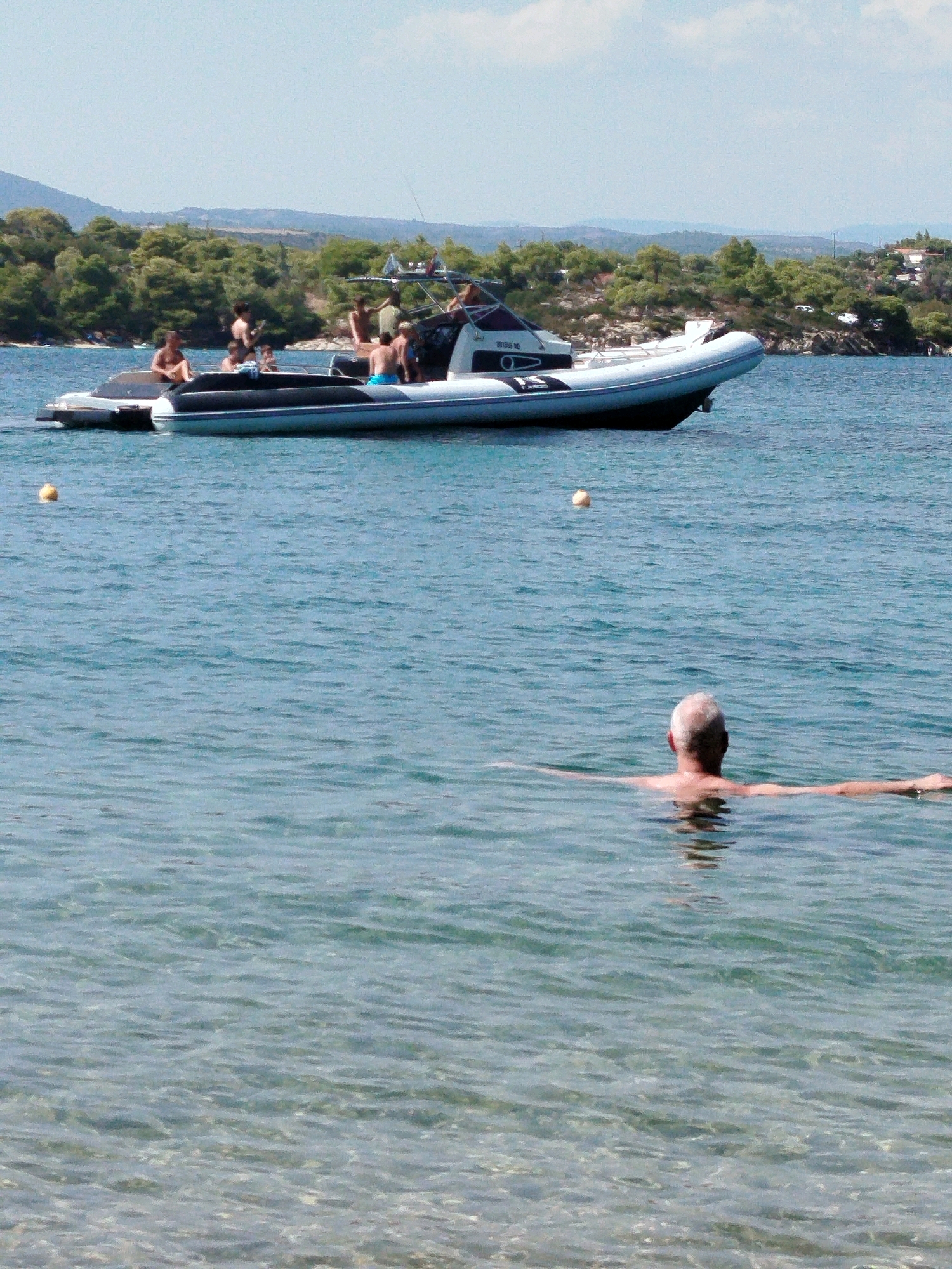 A wide shot captures a bright, sunny day at a beach in Sithonia Municipal Unit, Greece. In the foreground, a man with short, graying hair is swimming in clear, shallow water, his arms extended outwards. The water is calm and reflects the light. In the midground, a rigid inflatable boat (RIB) is moored, carrying several people. Some individuals are seated, while others appear to be standing or moving. The boat is white with dark trim and has a console with a windshield. In the background, a lush, green hillside covered in trees slopes down towards the sea, dotted with some buildings. The sky is a pale blue with a few wispy clouds. A yellow buoy is visible floating on the water to the left of the RIB.