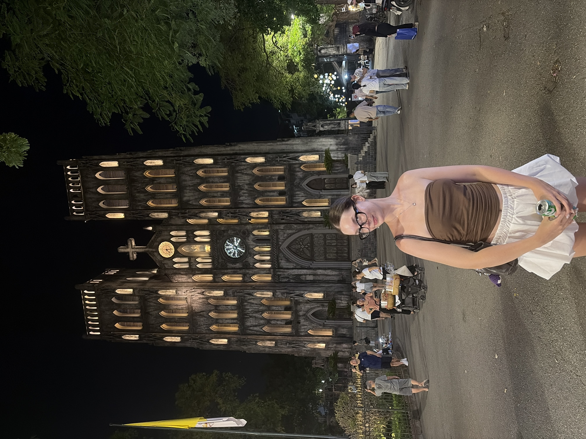 A young woman with glasses and brown hair is standing in front of Saint Joseph Cathedral in Hanoi, Vietnam. She is wearing a brown tube top and a white skirt. She is holding a can of soda. Behind her, the cathedral is illuminated at night. Several people are walking in the background. The cathedral, a neo-Gothic church, was built by the French colonial administration in 1886. It is a prominent landmark in Hanoi.