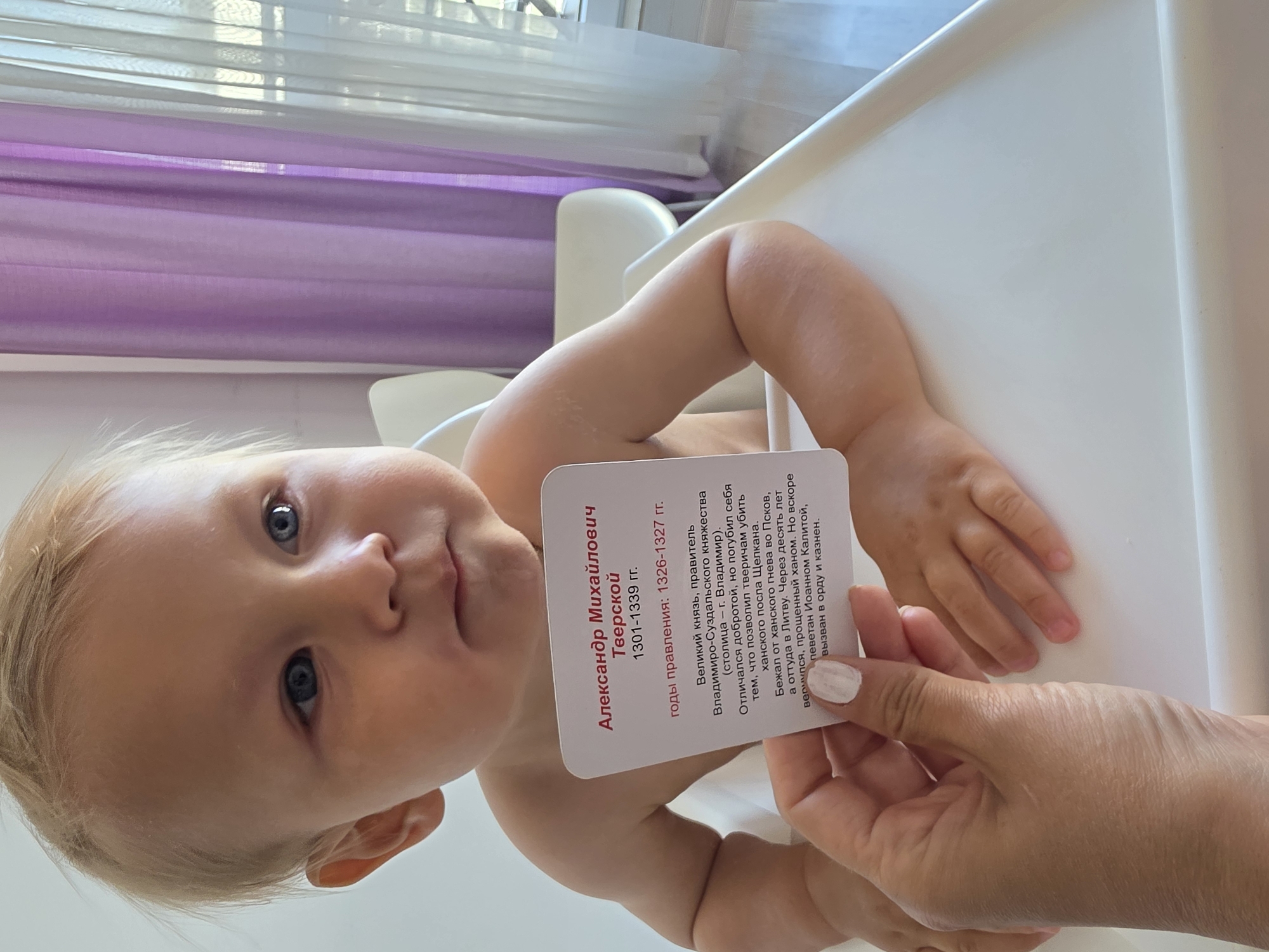 This media file features a close-up shot of a baby, likely a toddler, with fair hair and blue eyes, looking directly at the camera. The baby is propped up in a white high chair, with a blurred background of purple curtains and a window with blinds. A hand, with painted white fingernails, is holding a white card with text on it, positioned in front of the baby. The card appears to be a historical information card, with Cyrillic text visible, likely related to Russian history. The lighting suggests it is daytime. The overall scene conveys a domestic and potentially educational moment, where a child is being shown a card about a historical figure. The text on the card mentions "Александр Михайлович Тверской" and dates, suggesting it is a biographical note about a Russian Grand Prince. The location is identified as Sochi, Russia, though this context is not visually apparent in the image itself.