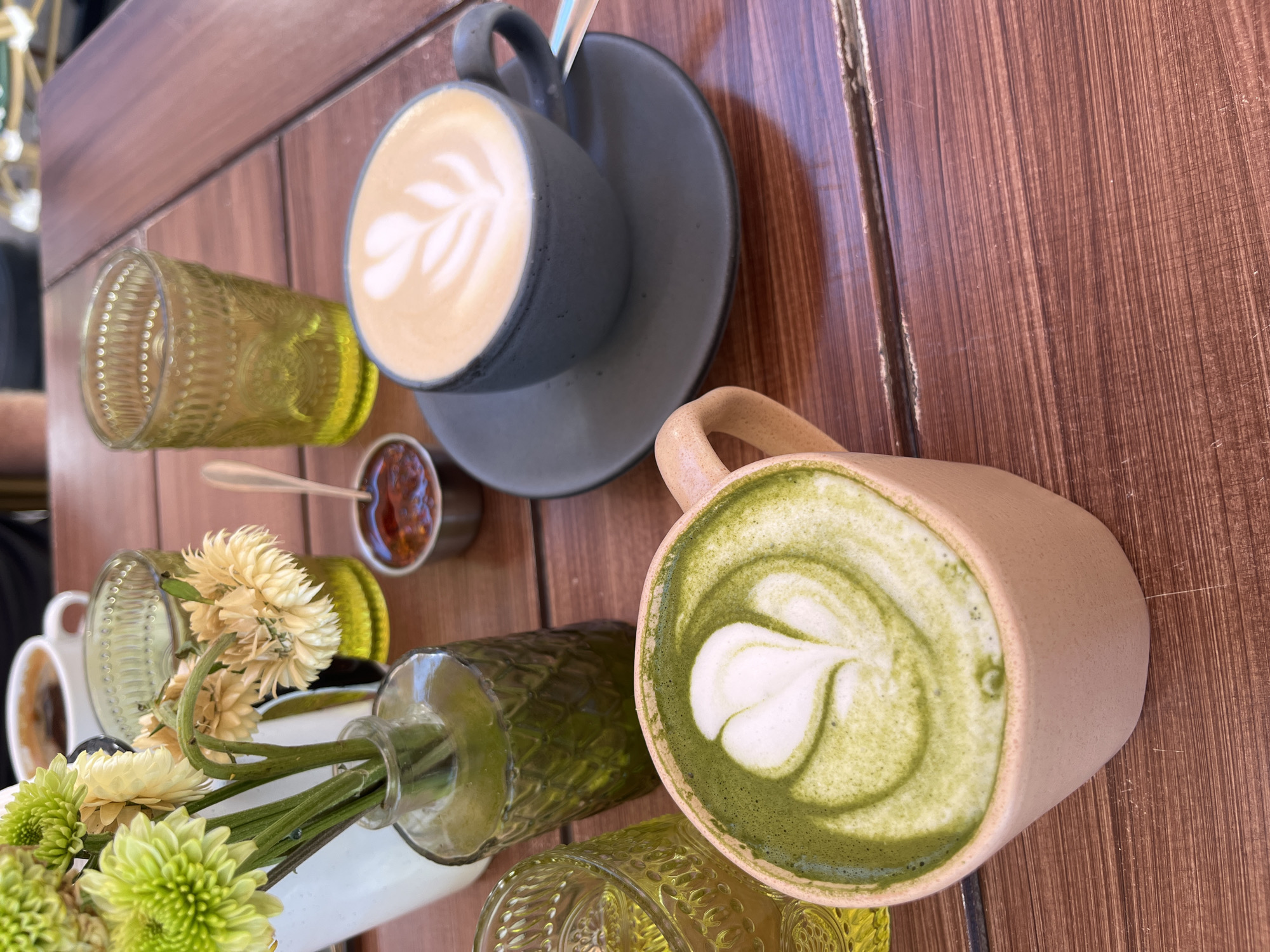 A high-angle shot on a wooden table features two cups of latte art, flowers in a vase, patterned glasses, and a small condiment container. One latte cup is gray, with a light-colored latte art design; it rests on a gray saucer. The other cup is beige, filled with a green matcha latte and a white latte art design. A small glass vase holding flowers is positioned near the matcha latte cup, and two patterned glasses are placed around the vase. A tiny dish of reddish condiment sits between the two latte cups. The scene appears to be set in San Bernardino Tlaxcalancingo, Mexico.