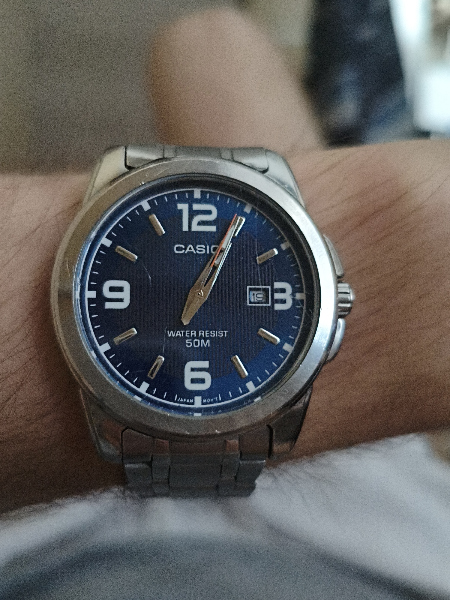 A close-up shot of a stainless steel Casio analog watch with a blue dial is worn on a person's wrist. The watch face displays white Arabic numerals for 12, 3, 6, and 9. The hour hand is pointing slightly past the 4, and the minute hand is pointing slightly past the 1. The second hand is a thin orange-tipped hand pointing at the 5. The watch also features a date window showing the 19th day of the month. The dial is textured with vertical lines and the text "CASIO", "WATER RESIST 50M", and "JAPAN MOV'T" are visible. The watch has a metallic band. The background is blurred and appears to be the person's arm and clothing, with some indistinct shapes in the distance. The image is taken from a high-angle perspective.