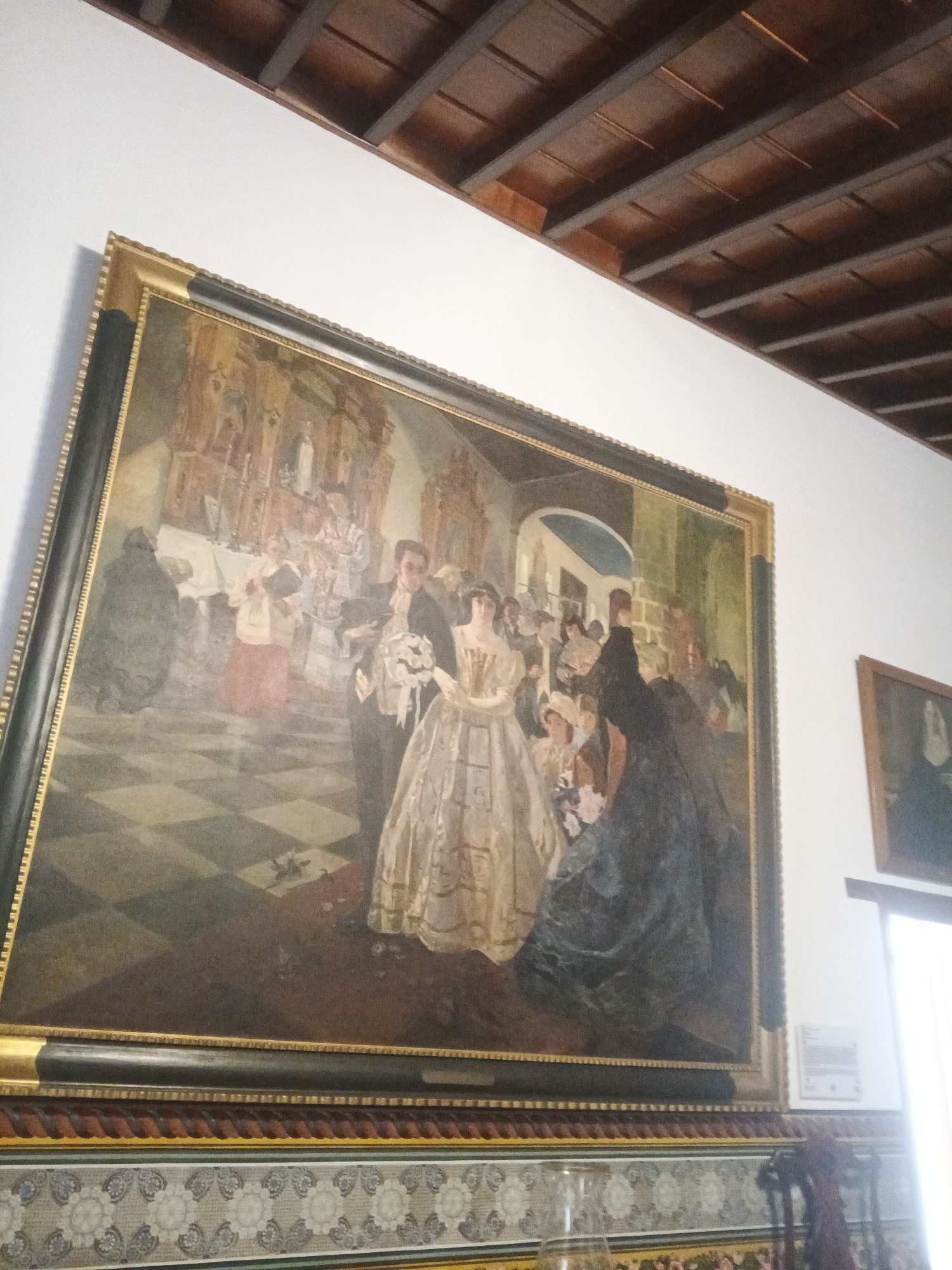 The image depicts a large, framed painting hanging on a wall. The painting appears to be a historical scene, showing a wedding in what seems to be a church or grand hall. In the foreground, a couple is prominently featured, likely the bride and groom, surrounded by other people in period clothing. The backdrop includes a checkerboard floor, an altar, and numerous figures attending the event. The painting is framed with a gold and black frame and hangs above a wall decorated with a floral pattern. In the background, a section of the ceiling is visible, made of wood beams. The exact location and city are not determinable from the image alone.