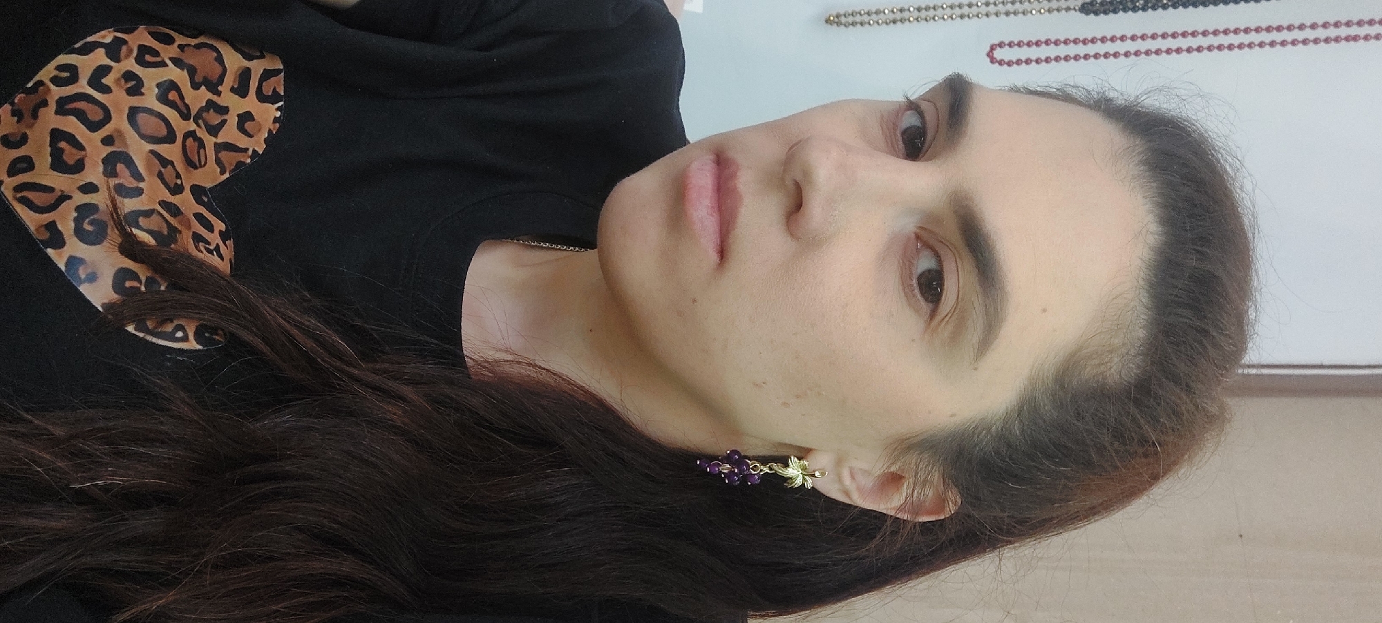 A woman is pictured indoors, facing the camera at an angle. She has long, dark brown hair and is wearing an earring shaped like a bunch of grapes on her right ear. She wears a black t-shirt with a leopard print heart on it. On the wall behind her are three necklaces, with beads of gold, black, and red. The exact location in Novosibirsk, Russia, cannot be determined from the image.