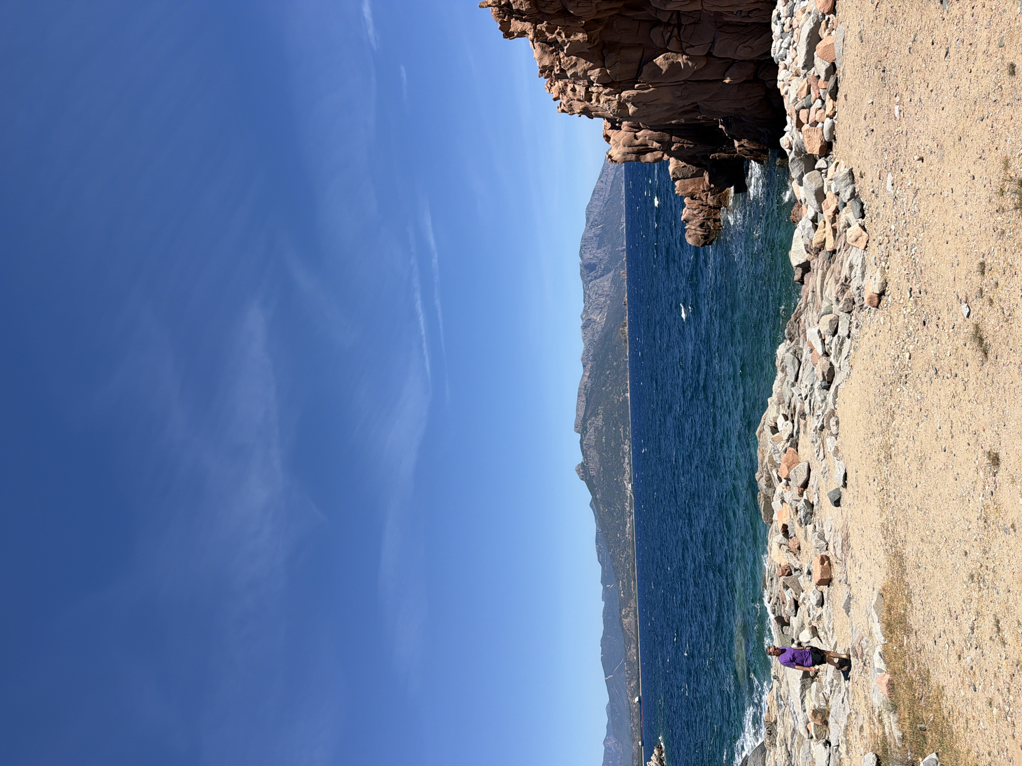 The image depicts a coastal landscape with a person walking along a rocky shore. The person, wearing a purple shirt and dark shorts, is positioned on the left side of the frame, on a path made of sand and scattered rocks. The sea, with its deep blue water and visible waves, stretches out to the horizon. In the background, a mountainous coastline is visible under a clear blue sky with some wispy clouds. The right side of the image is dominated by a large formation of reddish-brown rocks. The scene appears to be a scenic natural area, likely a public park or a hiking trail along the coast of Tortolì, Italy, known for its beautiful Mediterranean beaches and rugged terrain.