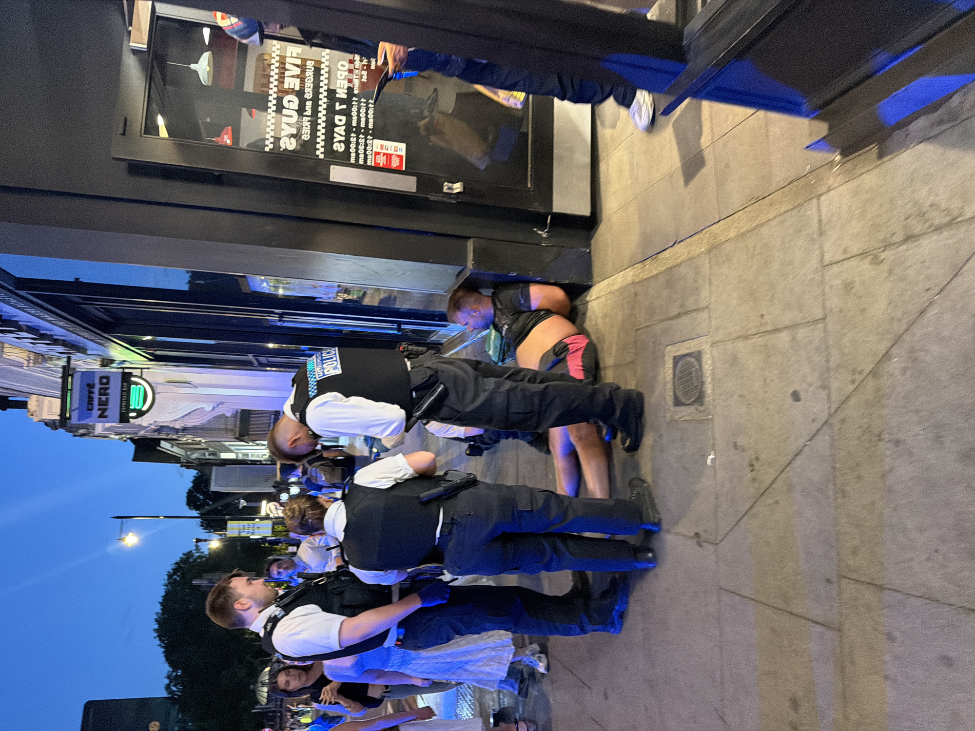 The scene depicts three police officers attending to an individual who is crouched or lying on the sidewalk. The officers are wearing police uniforms with vests labeled "POLICE". One is kneeling and appears to be examining or assisting the individual. A crowd of onlookers stands nearby, some holding mobile phones, suggesting they may be recording the event. The incident is occurring outside a "FIVE GUYS" restaurant and a "CAFFÈ NERO" in an urban environment, likely the City of London, United Kingdom. The presence of streetlights and the evening sky indicate that the photo was taken at night.