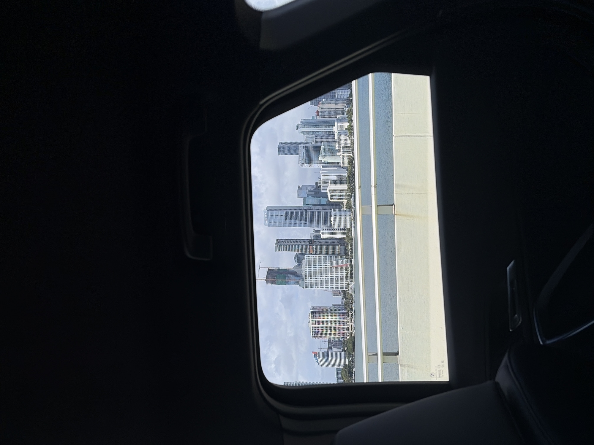 The image shows a view of the Miami skyline, as seen through an open sunroof of a vehicle. The buildings are varied in height and design, typical of an urban environment. There are some cranes visible on the skyline, which suggests ongoing construction. The skyline is viewed through the open sunroof, indicating the vantage point is from inside a car. The background sky is lightly overcast, suggesting a cloudy day. The vehicle interior is dark.