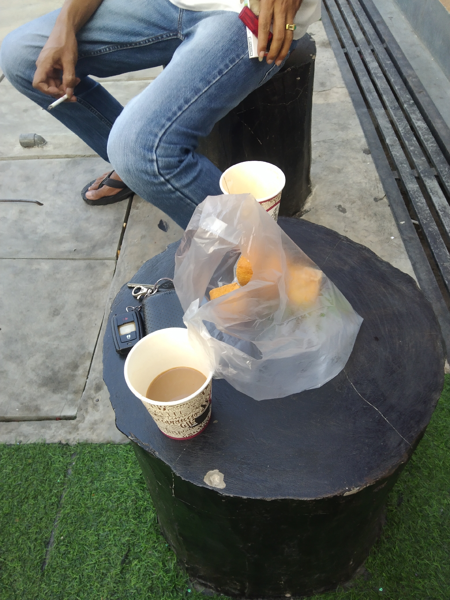 A person is sitting outside, holding a cigarette and a lighter, with a cup of coffee and a bag of food on a dark, circular table. The person is wearing blue jeans and a white shirt. They are also wearing a ring on their right hand. The table is made of wood and has a rough texture. There is also a set of keys and a car remote on the table. The environment is a public space with a concrete floor and a grassy area. A metal bench is visible in the background. The lighting suggests it is daytime. The image is cropped to focus on the table and the person's lower body. Based on the provided information, the location is in Surawangi, Indonesia.