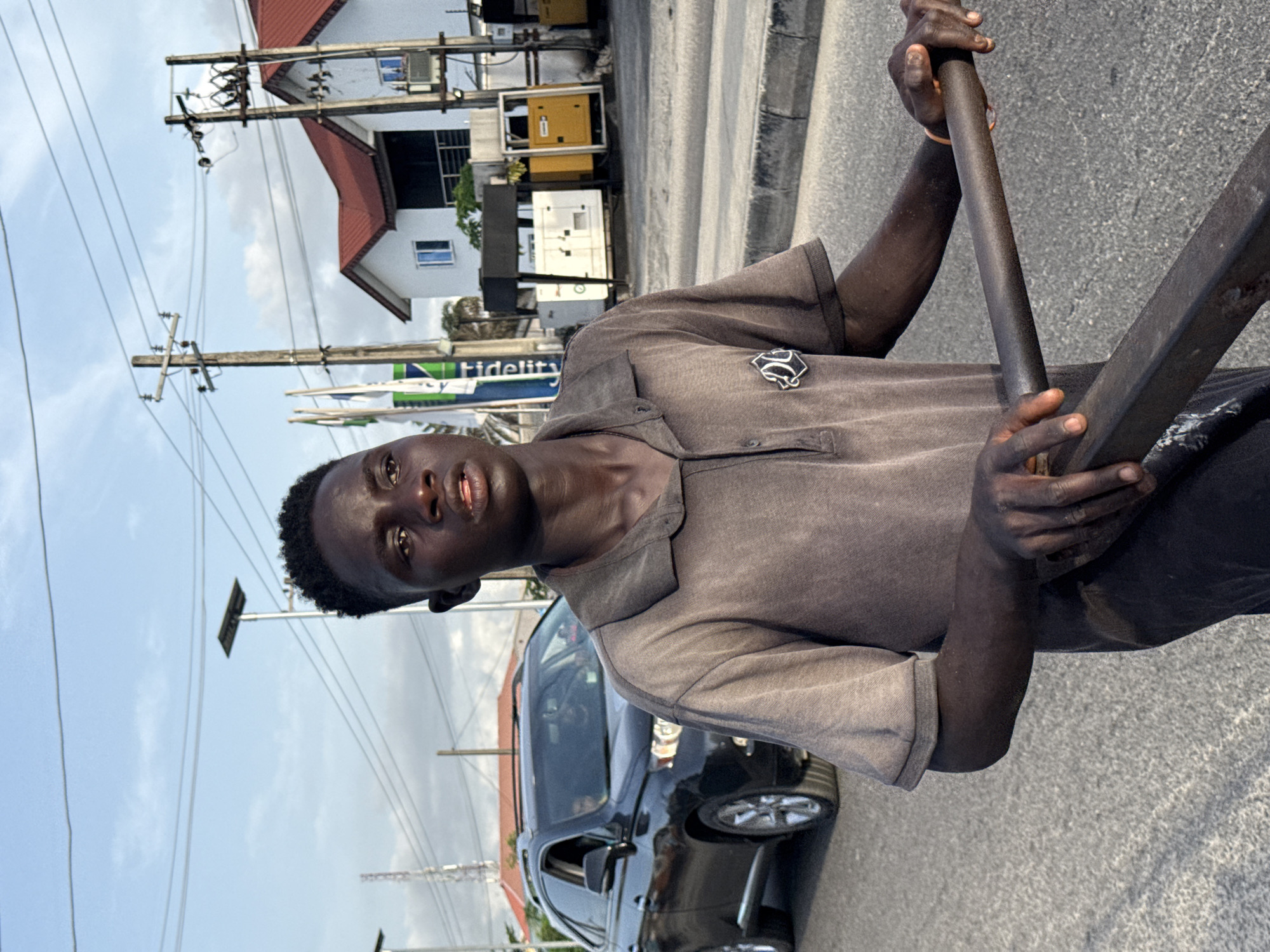 An individual with dark skin stands on a gray asphalt surface, holding a metal rod in their hands. They wear a gray collared shirt and dark pants. Behind the person is a dark-colored vehicle. In the background, there are buildings, including a white structure with a red roof, and electrical poles with numerous wires. A sign with "Fidelity" is also visible. The scene appears to be a street in Ado, Nigeria.