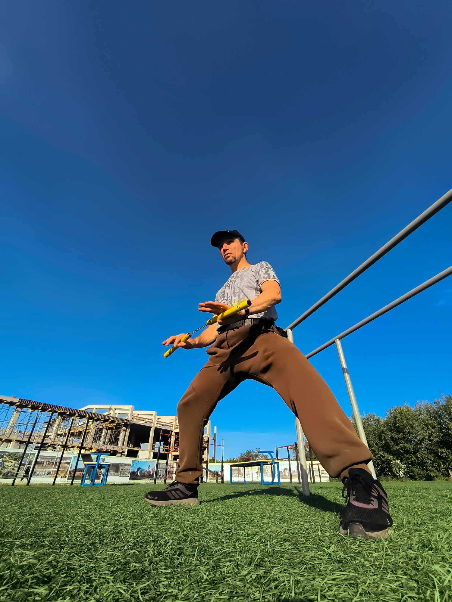 A man in a baseball cap, t-shirt, and brown sweatpants stands on a grassy field with a bright blue sky overhead. He is holding yellow nunchaku. Behind him are parallel bars, a construction site with scaffolding, trees, and a blue structure. The scene appears to be an outdoor sports or training area.