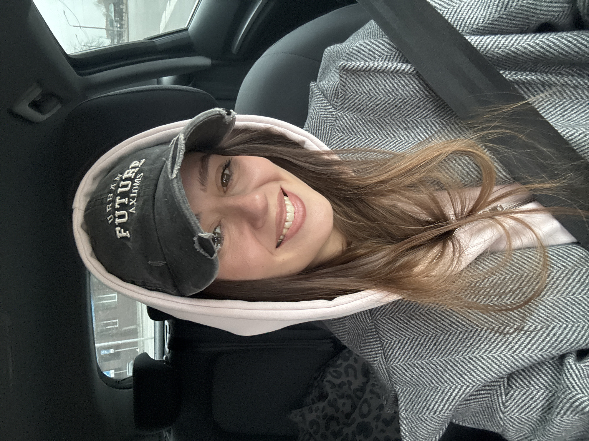 A woman wearing a baseball cap with the words "URBAN FUTURE AXIOMS" and a pink hoodie under a herringbone-patterned coat is sitting in a car. She is wearing a seatbelt and smiling at the camera. Through the car window, a building with windows can be seen. The photo appears to be taken in overcast or rainy weather. It is not possible to determine the specific location in Krasnoyarsk, Russia from the provided image.