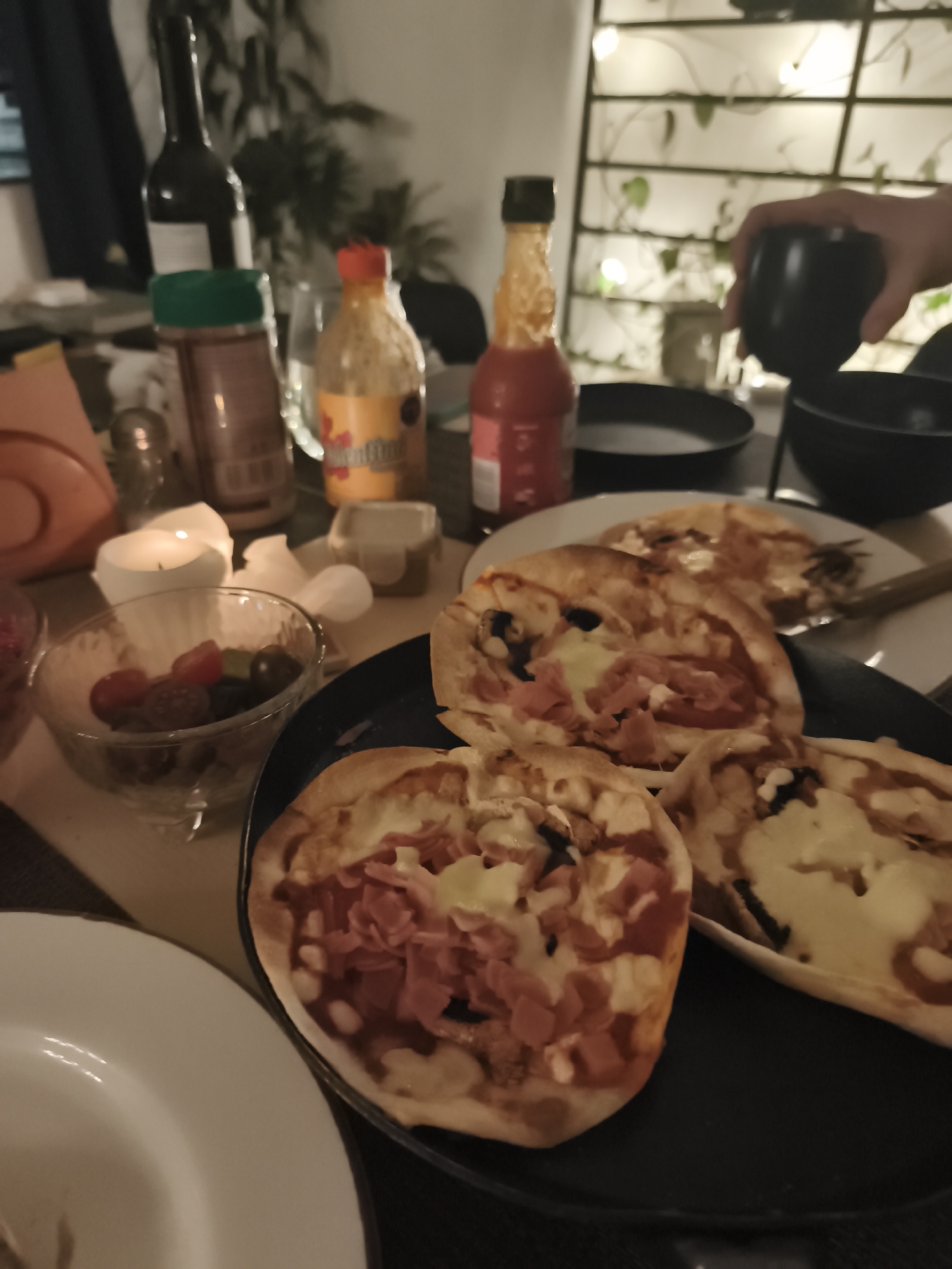 A nighttime indoor scene shows a table laden with food and drink. Two black plates hold small pizzas, some topped with ham and black olives, and others with melted cheese. A white plate is visible in the foreground. Nearby, a glass bowl contains a mix of red and green fruits. Behind the pizzas, there are several bottles, including a wine bottle, a spice jar, and two bottles of hot sauce. A white candle in a holder and a small container are also present. In the background, a metal trellis decorated with lights and green plants is visible. A hand is partially visible holding a black cup. The location is assumed to be in Mexico City, Mexico.