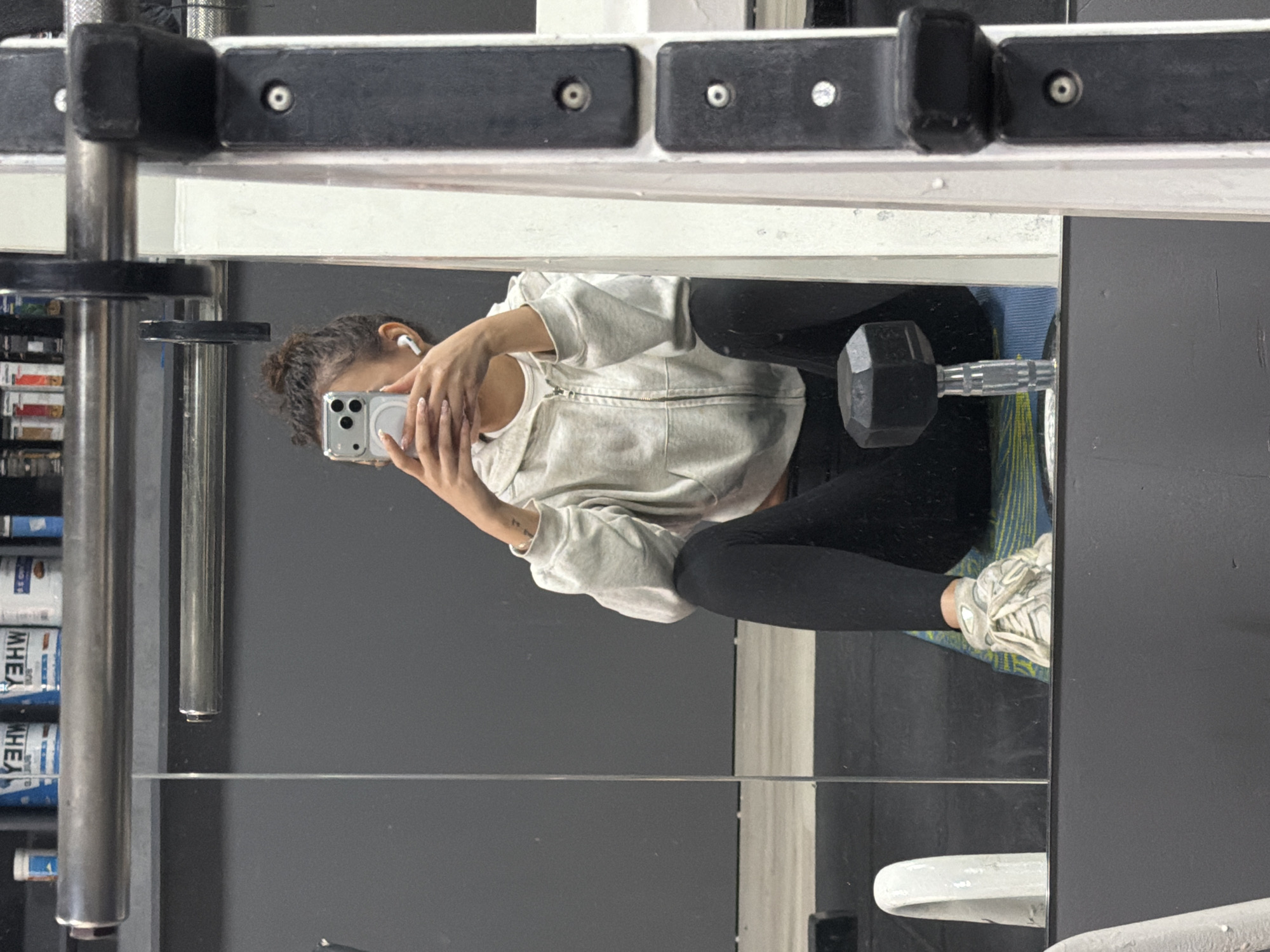 A person is sitting on the floor in a gym, visible in a mirror. The person is wearing black leggings, a light-colored zip-up hoodie, and white sneakers. They are holding a smartphone in their hands, presumably taking a photo or video of themself in the mirror. The location appears to be a gym in Sofia, Bulgaria. Gym equipment such as a weight rack, dumbbells, and other fitness accessories are visible in the reflection. The environment includes a grey wall and a blue and yellow workout mat.