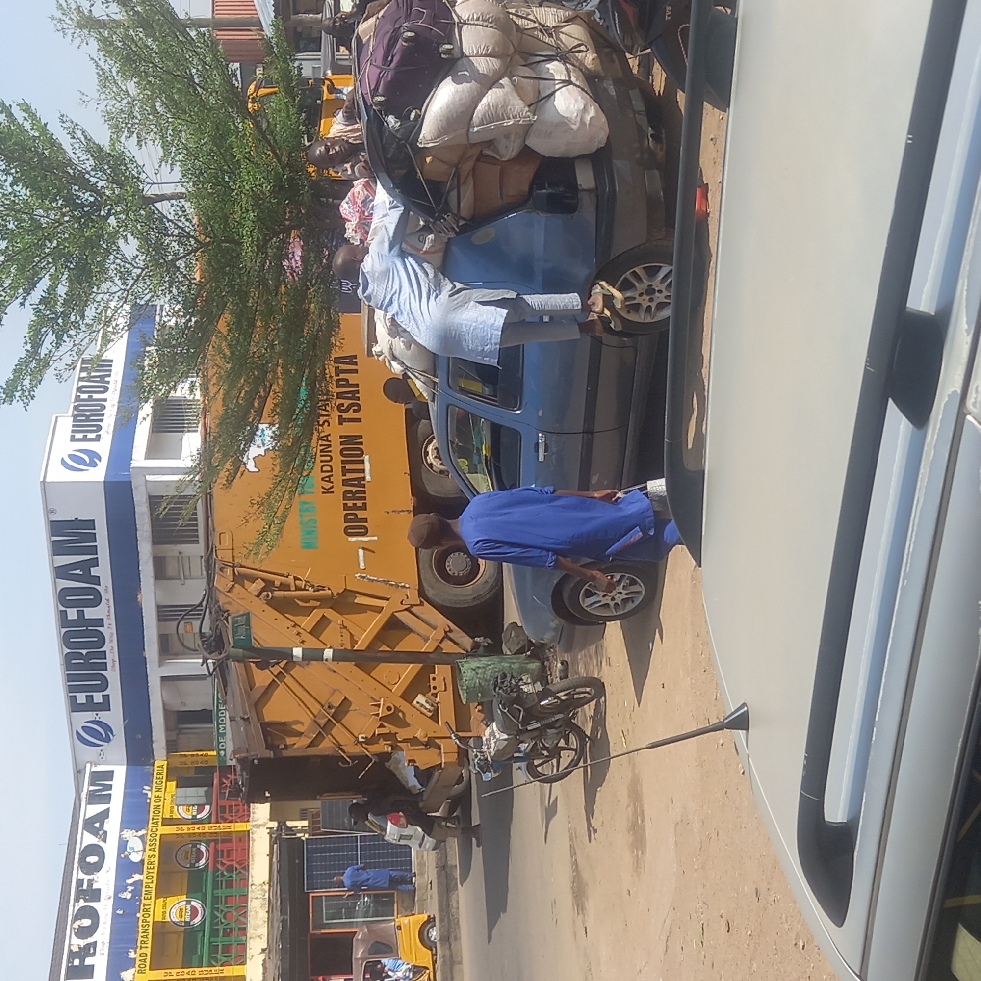 The image depicts a street scene in Dadi Riba, Nigeria, with a blue car heavily loaded with goods and people on top. A man in a blue robe is standing next to the car. Behind the car, there is a yellow truck with the words "KADUNA STATE OPERATION TSAPTA." Adjacent to the truck is a building with a sign that reads "ROFOAM EUROFOAM," and another sign for the "ROAD TRANSPORT EMPLOYER'S ASSOCIATION OF NIGERIA." There is also a motorcycle parked near the truck. The image also shows other people and vehicles, including a white car, in the surrounding area.