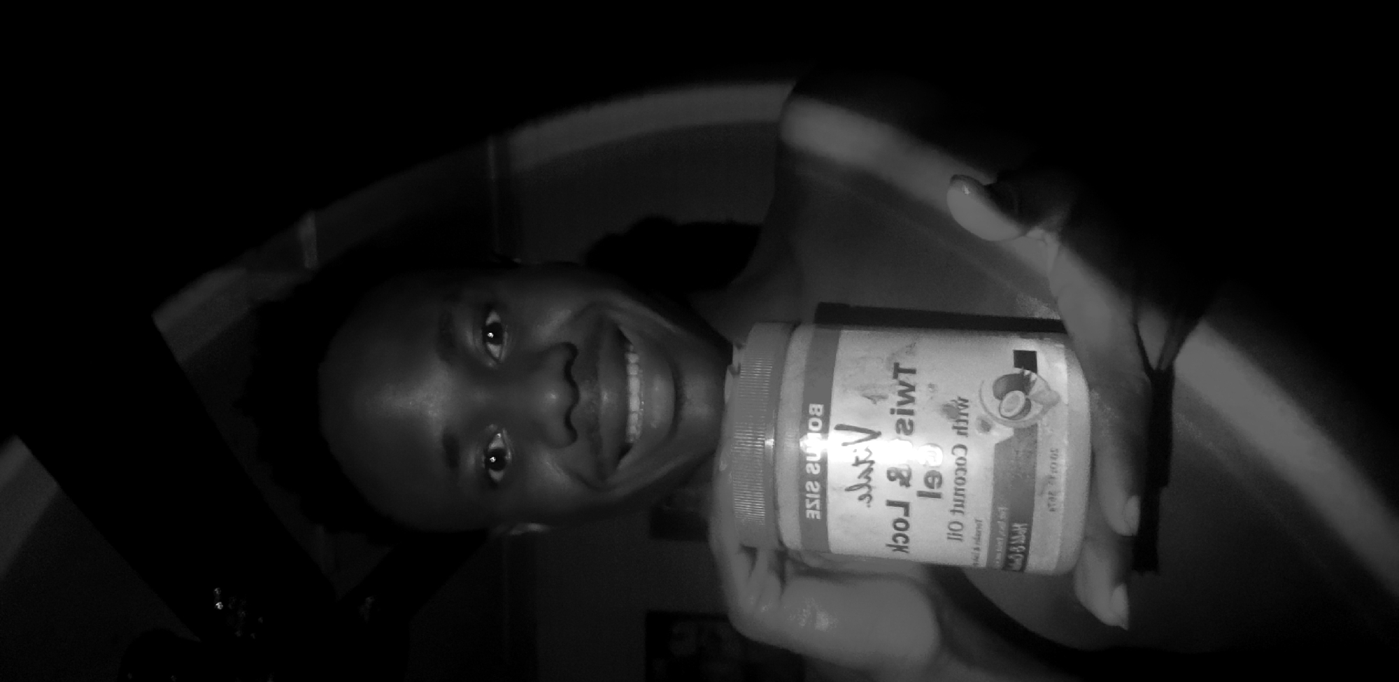 A close-up, black and white, chest-up shot shows a young Black man with short, textured hair, holding a jar of hair product. He is smiling broadly, with his teeth showing. The jar is white with a red lid and a label featuring text and an image of a coconut. The lighting is dramatic, with strong shadows obscuring parts of the background and the subject's body. It is unclear if this image was taken in Narayi, Nigeria.
