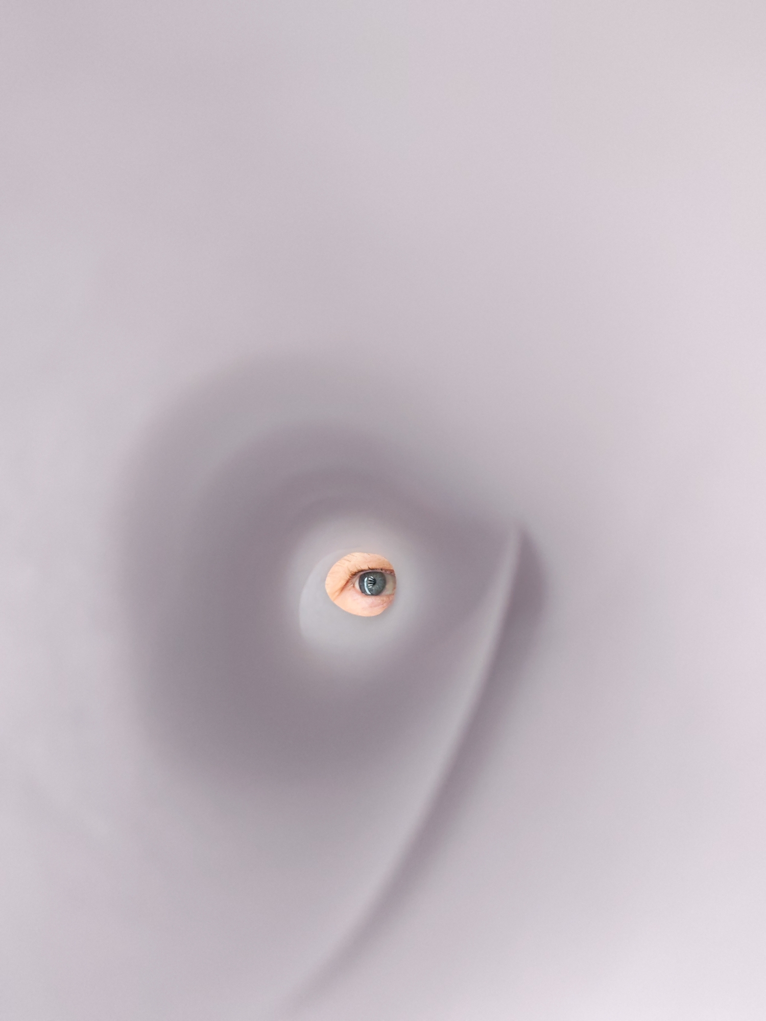 The image features a single human eye peering through a circular hole within what appears to be a rolled sheet of light grey or white paper. The eye is light-colored, possibly blue or green, and the skin around the eye is visible. The paper is the dominant element, creating a blurred tunnel effect that draws focus to the eye. The background is indistinct. The exact location in the city of Chaikovsky, Russia is not discernible from the image.
