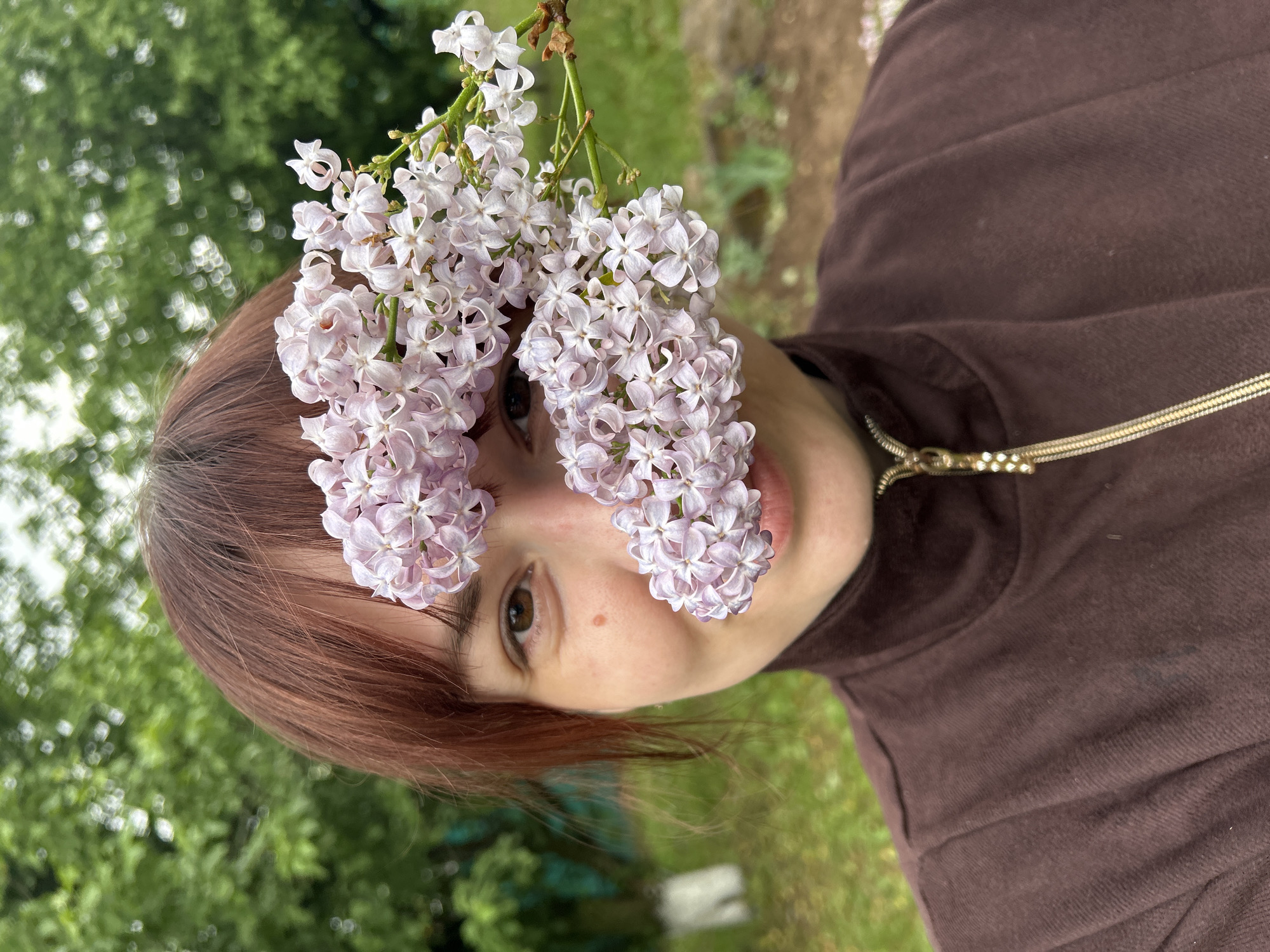 A person with red hair and a brown top is holding two bunches of lilac flowers in front of their face. The background shows green trees and grass. The location is not discernible.