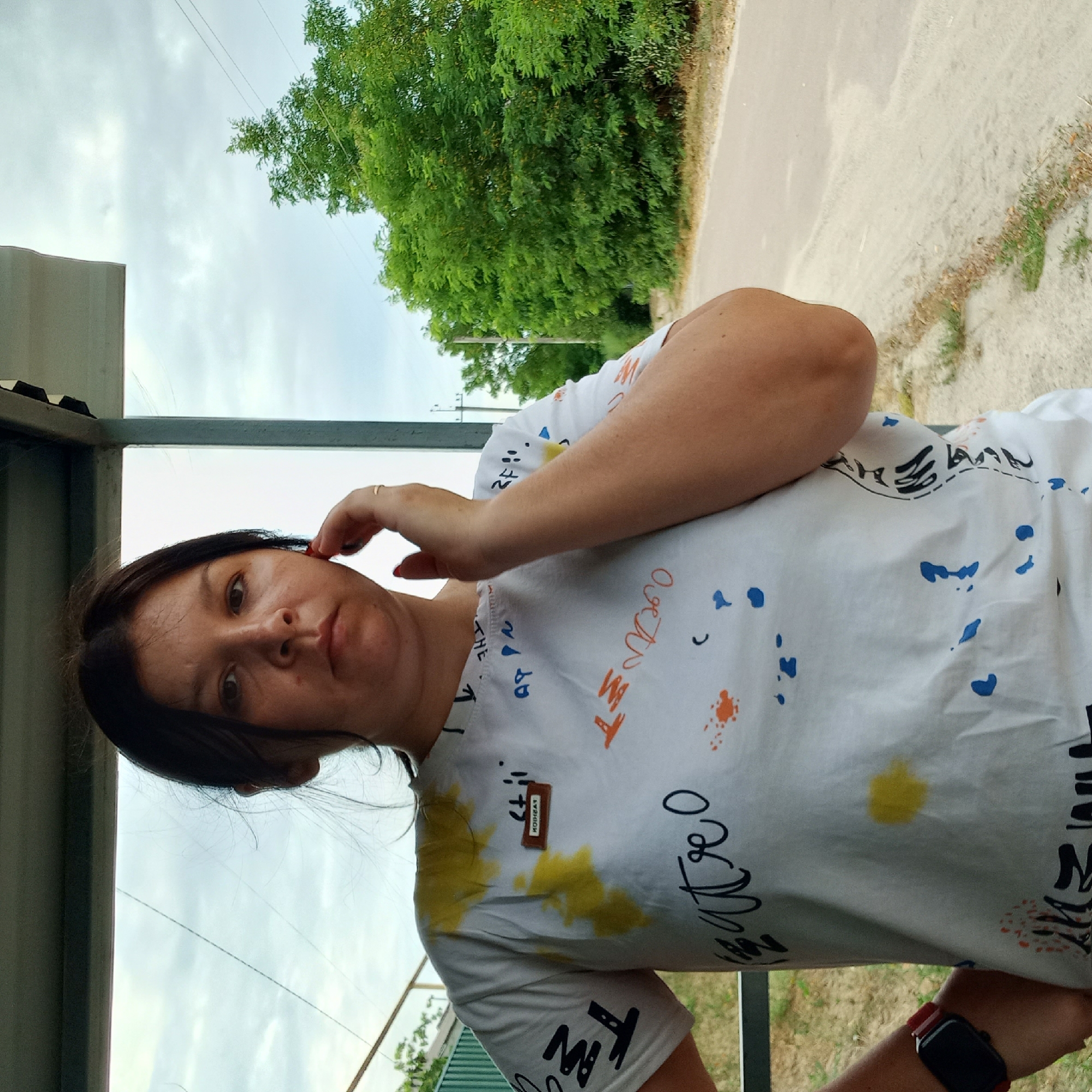 A young woman with dark hair and brown eyes is standing in an outdoor shelter, possibly a bus stop or covered seating area. She is wearing a white t-shirt with a colorful, abstract design and text. Her right hand is raised to her ear, and she is wearing a ring on her right ring finger. She also has a smartwatch with a red strap on her left wrist. Behind her, a road is visible, bordered by green trees and vegetation under a cloudy sky. The overall scene appears to be a casual, outdoor setting.