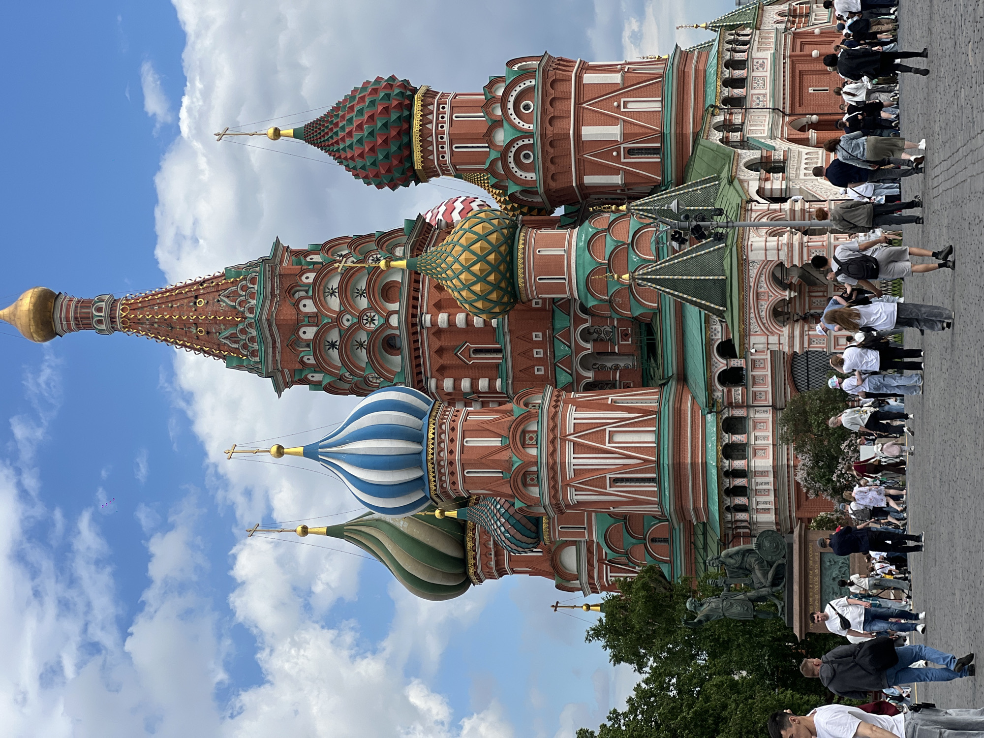 The image shows a vertical view of Saint Basil's Cathedral in Moscow, Russia, with a crowd of people in the foreground. The cathedral features multiple colorful onion domes, distinctive architectural details, and a red brick facade. The domes are patterned with various colors and designs, including stripes, scales, and diamond shapes. A monument depicting figures is visible in front of the cathedral. People are walking and standing near the monument. The sky above the cathedral has blue sky and white clouds.