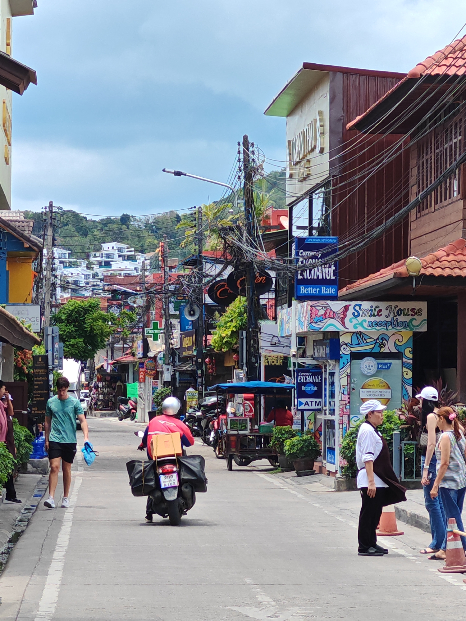 The image shows a street scene in Baan Bo Phut, Thailand, with a narrow road lined by buildings on both sides. The architecture is a mix of traditional Thai styles with wooden elements and more modern structures. Overhead, a dense network of electrical wires crisscrosses the sky, a common sight in Southeast Asia. Several signs are visible, indicating businesses like "Currency Exchange," "ATM," and "Smile House reception."
In the foreground, a young man wearing a turquoise t-shirt and black shorts walks on the left side of the street, carrying a blue bag. To his right, a person on a motorbike is driving away from the camera. The motorbike has a large box strapped to the back, and the rider is wearing a red jacket and a silver helmet.
Further down the street, there are more shops and stalls, some with displays of goods. A few people are visible standing on the sidewalk, including a woman in a white chef's uniform and hat and two other women. The background reveals a hilly landscape covered in lush greenery, with white buildings dotted along the slopes, suggesting a resort or residential area. The sky is partly cloudy. The overall impression is of a bustling, vibrant street in a tropical location.