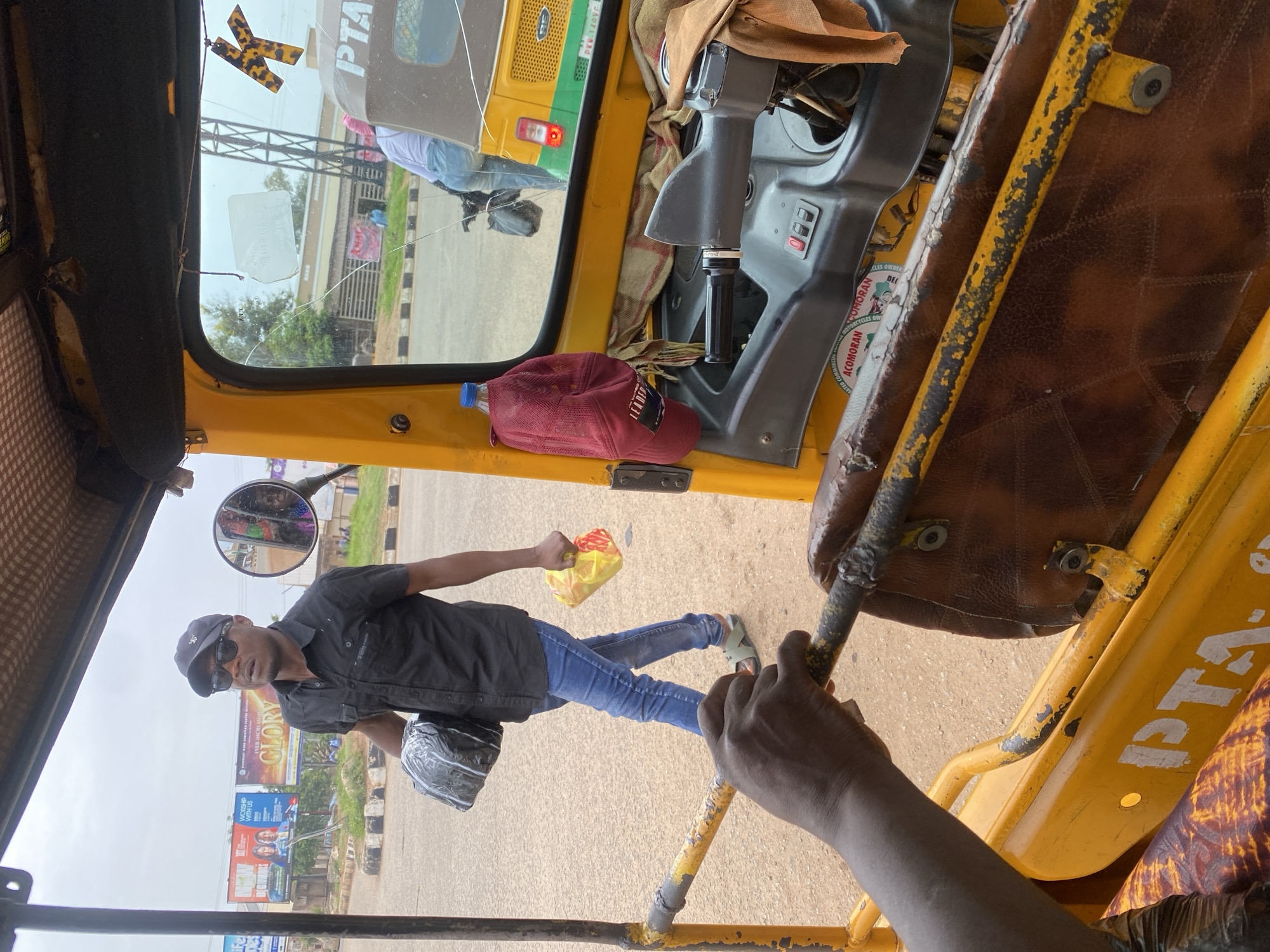 In Jos, Nigeria, a man wearing a black short-sleeved shirt, blue jeans, sunglasses, and a baseball cap walks down a street carrying a black package in his left arm and a yellow plastic bag in his right hand. He is walking past a yellow and green auto-rickshaw parked on the side of the road. The interior of the auto-rickshaw is visible, showing a brown seat, a steering wheel, and a dashboard with red buttons. A red baseball cap hangs from a hook inside the auto-rickshaw, and a blue water bottle is also visible. In the background, billboards and other vehicles are present, suggesting an urban environment. The sky is overcast.