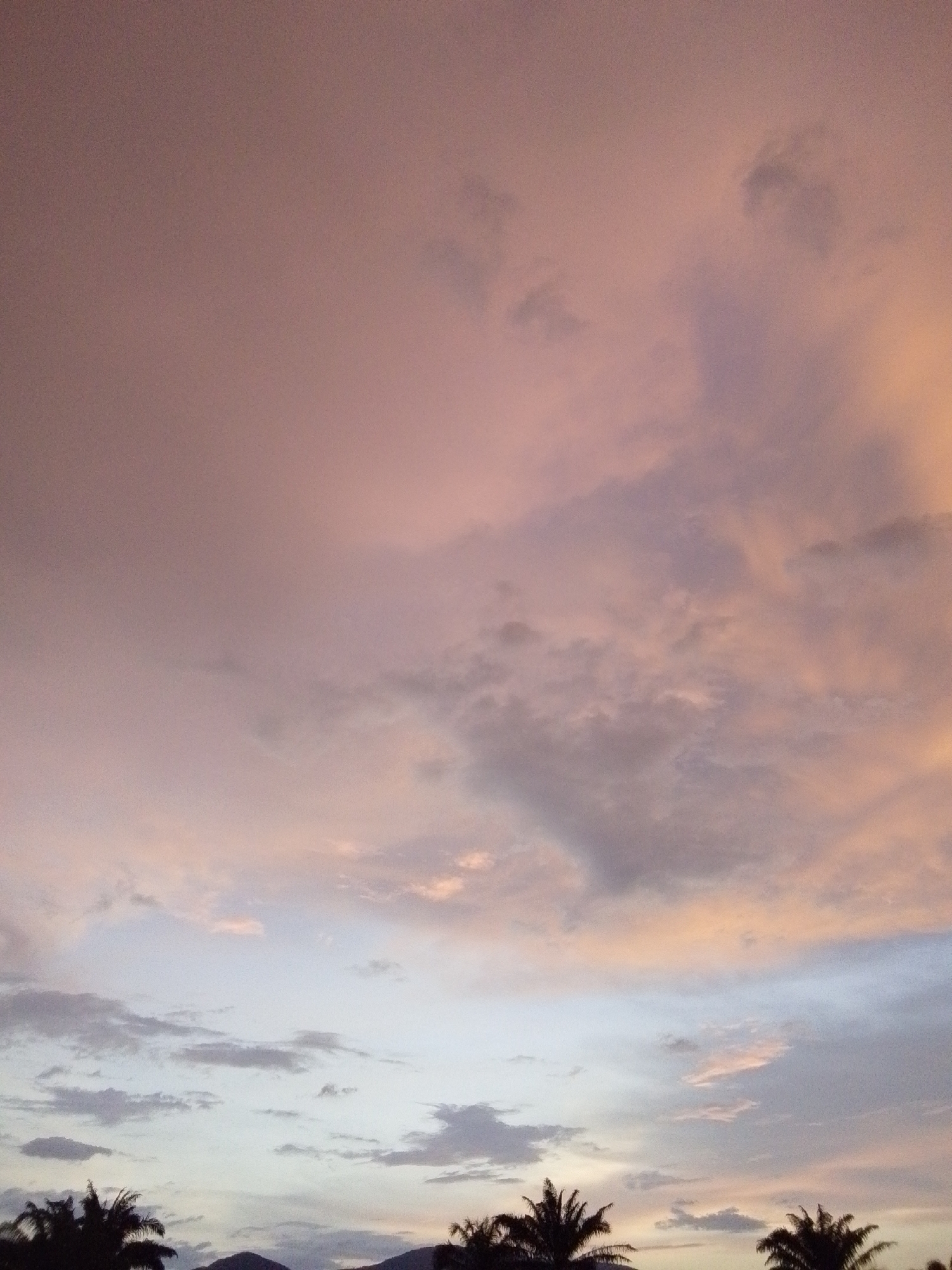 The image captures a sky filled with clouds, exhibiting hues of pink, orange, and gray, characteristic of a sunset or sunrise. Palm trees are silhouetted against the lower portion of the sky, with faint outlines of hills or mountains visible in the distance. The scene appears to be an outdoor environment, likely a tropical or subtropical region, possibly in or around Serti, Nigeria. There are no discernible people, specific objects, or actions taking place within the frame.
