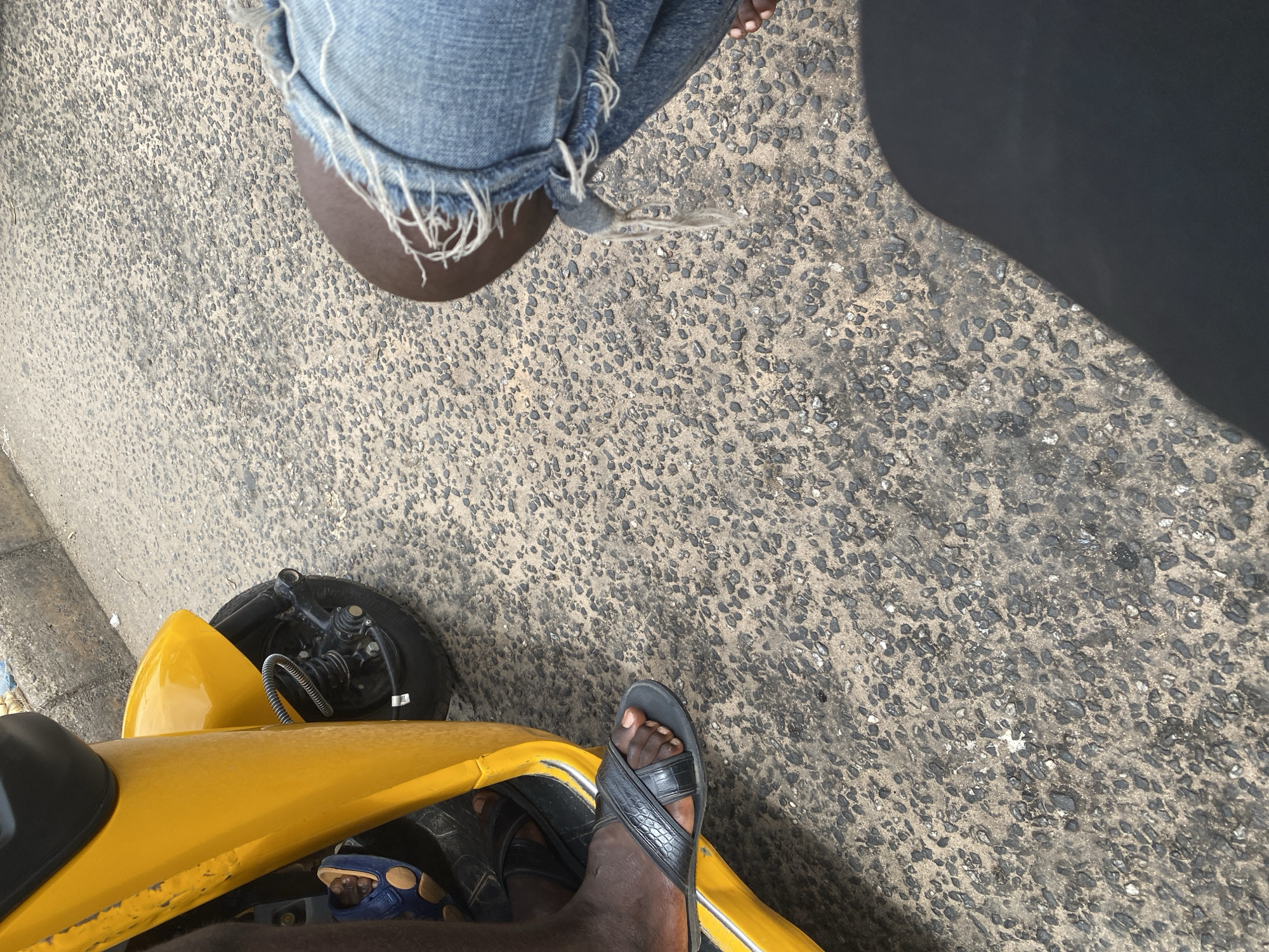 The image captures a low-angle perspective from a yellow vehicle, likely a motorcycle taxi or tricycle, in Jalingo, Nigeria. The foreground shows a person's feet wearing black and blue sandals, resting on the vehicle. The person is wearing ripped denim jeans, with frayed edges visible. The ground is a textured asphalt surface, typical of roads. A large, dark shadow falls across the right side of the image, suggesting it is a sunny day. The background includes the wheel and mechanical components of the vehicle, as well as a glimpse of a sidewalk or curb on the left. The scene appears to be outdoors, on a road, possibly during daylight hours. There is no visible text in the image.