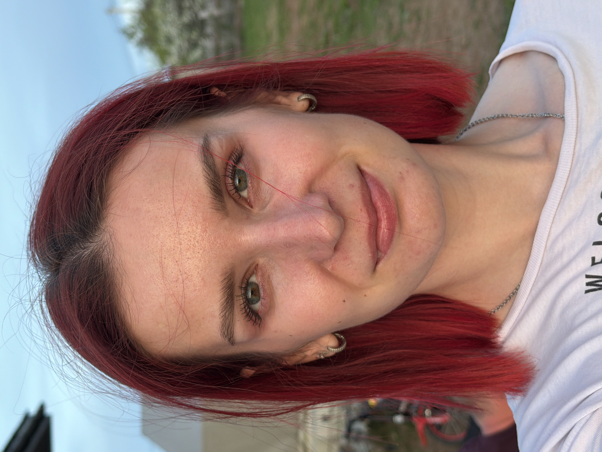 A portrait of a young woman with dyed red hair, light skin, and green eyes. She is wearing a white t-shirt and a silver necklace. She has earrings in her ears. The background appears to be an outdoor setting, possibly a park or garden, with glimpses of greenery and a blurred skyline. There is no information about the location being in the city of Chaikovsky, Russia in this image.