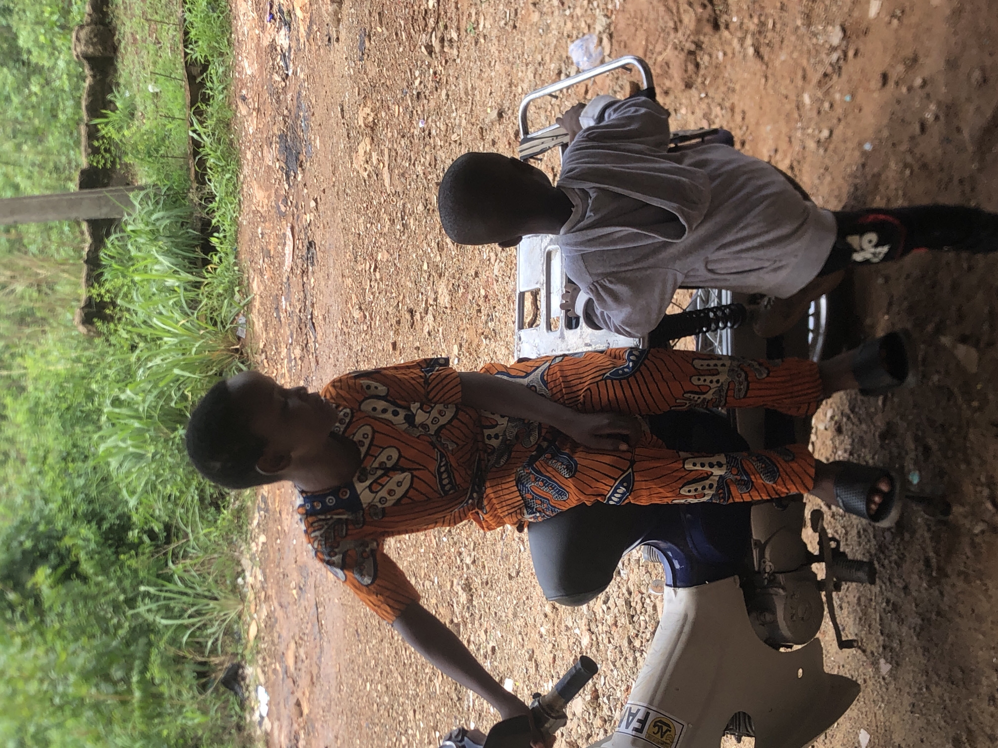 Two young individuals are present in this outdoor scene in Nkwelle, Nigeria. One person is sitting on a stationary motorbike, while the other is positioned behind them, possibly interacting with the bike's rear carrier. The individual on the bike is wearing an orange and brown patterned garment, and sandals. The person in the rear is dressed in a gray long-sleeved shirt and dark-colored bottoms. The setting appears to be a rural or semi-rural area in Nkwelle, with a dirt road and vegetation visible.