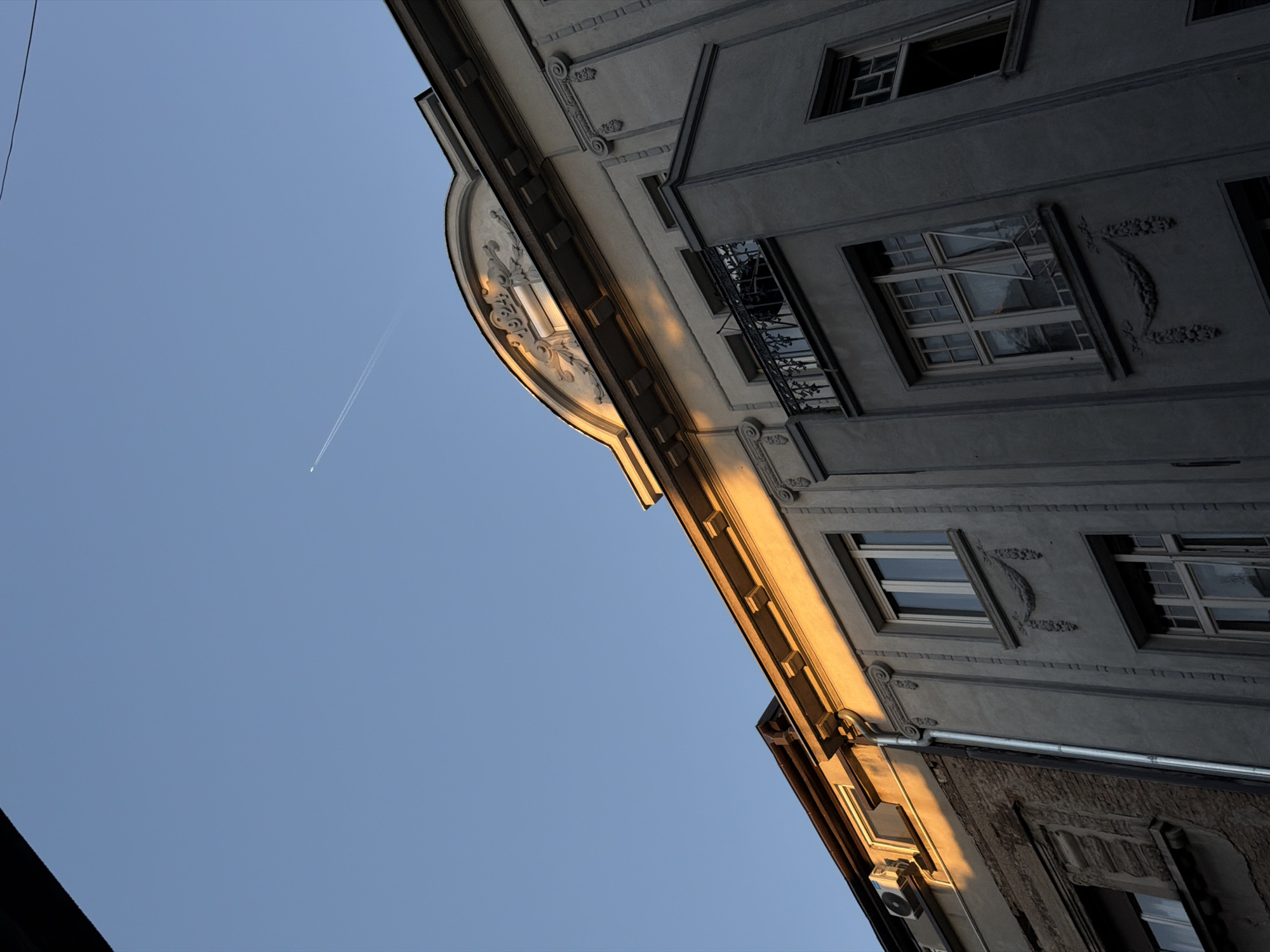 A low-angle shot captures the architecture of a building in the Vracar Urban Municipality, Serbia. The building's facade features ornate detailing, windows with open panes, and a balcony with a black wrought-iron railing. A section of the building is illuminated by sunlight, creating a warm glow on the stone. Above the building, a blue sky is visible with a contrail from an airplane. Power lines are visible in the upper left corner of the frame.