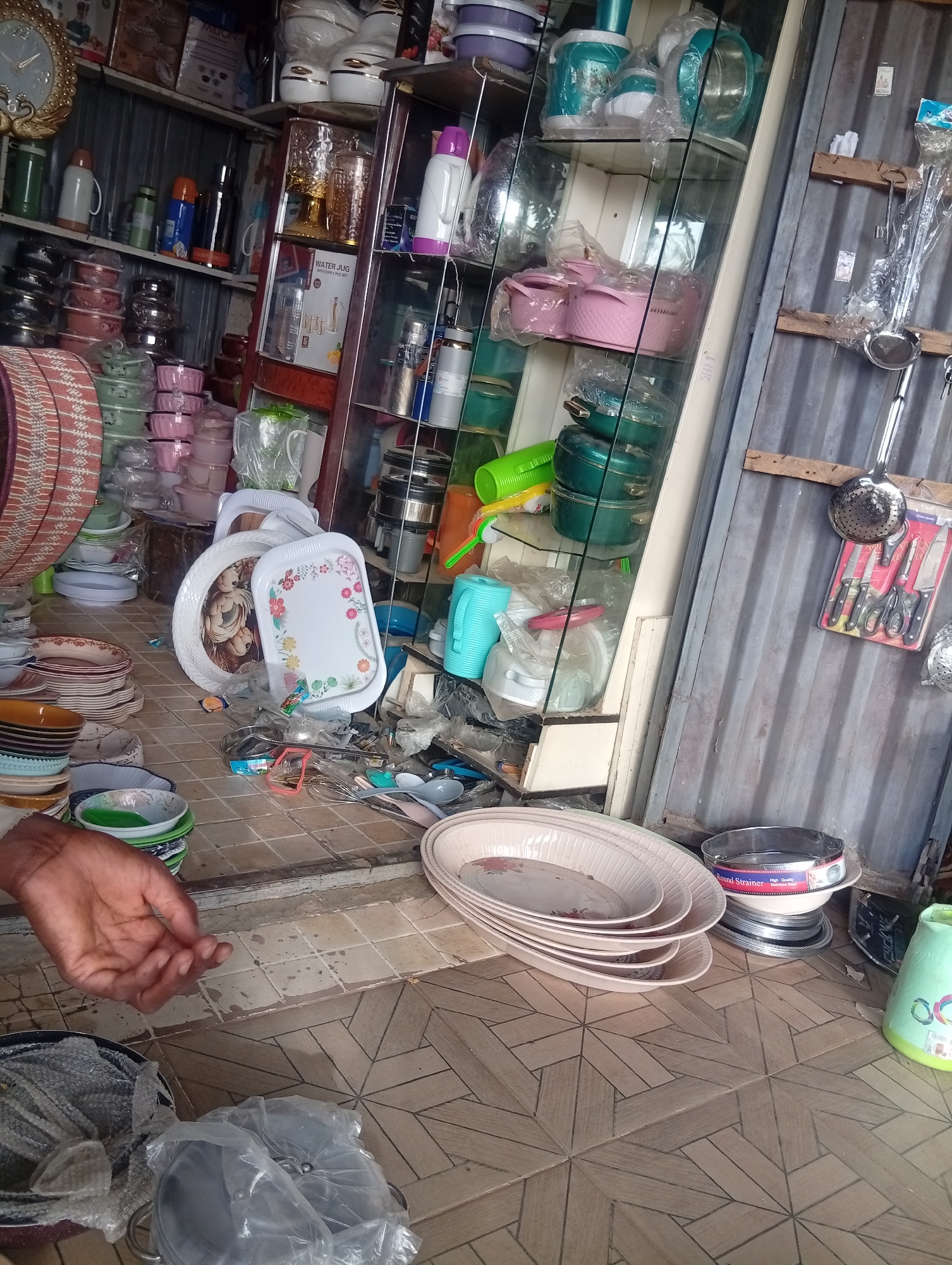 This image captures a densely packed stall or small shop specializing in kitchenware and household goods, likely in Birnin Kudu, Nigeria. The setting appears to be a retail space, possibly semi-outdoor, with corrugated metal sheets forming a wall on the right and a geometrically patterned tiled floor.
In the foreground, a person's dark-skinned hand is visible on the lower left, resting near stacks of colorful plates and bowls. Adjacent to this, various items are displayed on the floor, including a green plastic basket, several light pink oval serving trays with floral designs, and a round metal strainer labeled "Round Strainer" and "High Quality Stainless Steel." Pots and pans, some wrapped in plastic, are also scattered on the floor.
The shop is overflowing with merchandise arranged on shelves and within a glass display cabinet. On the left, shelves hold an assortment of thermos flasks in green, white, blue, orange, and black/silver, alongside stacks of bowls in pink, green, and purple hues, and pink cooking pots. An ornate golden clock is visible on the upper left wall, and a box bearing the brand "MILTON" is on a higher shelf.
The central glass cabinet features glassware, teal and pink cooking pot sets, water jugs, and decorative serving trays, one with a floral pattern and another with a teddy bear design. A box inside the cabinet clearly reads "WATER JUG" and "6PCS SET." To the far right, hanging on the corrugated metal wall, are various kitchen utensils, including a packaged set of knives and scissors, a metal ladle, and a metal skimmer. The scene suggests a busy commercial environment filled with household necessities.