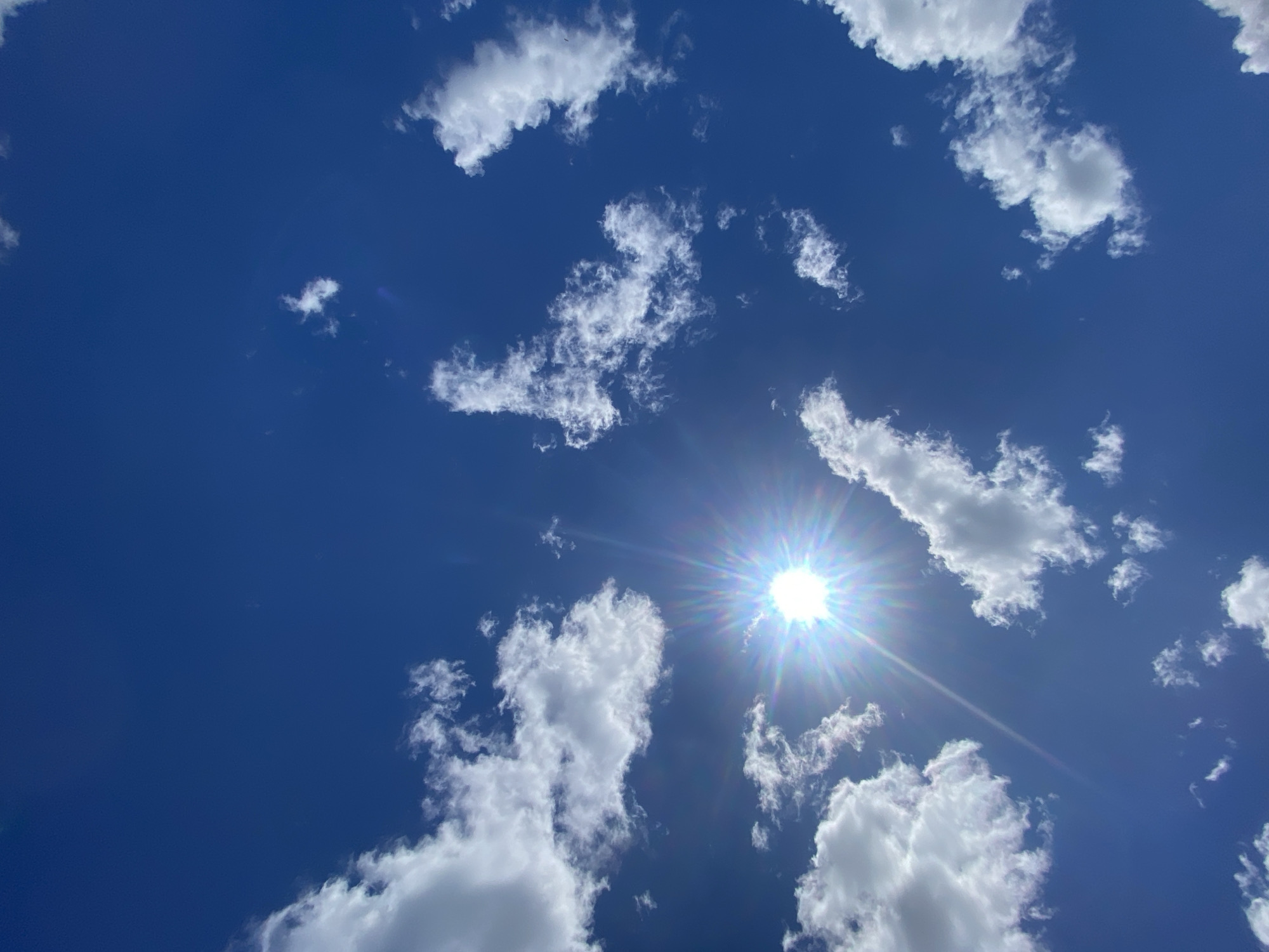 The image depicts a bright, sunny day with a clear blue sky dotted with white, fluffy cumulus clouds. The sun is prominently visible, shining brightly and creating a lens flare with distinct rays of light emanating outwards. The perspective is looking directly up at the sky, suggesting the photo was taken outdoors in an open area. The scene is serene and natural, with no man-made structures or individuals visible. The environment is characteristic of fair weather, likely during daylight hours. No specific location in Ustyvytsia, Ukraine can be identified from the image alone.