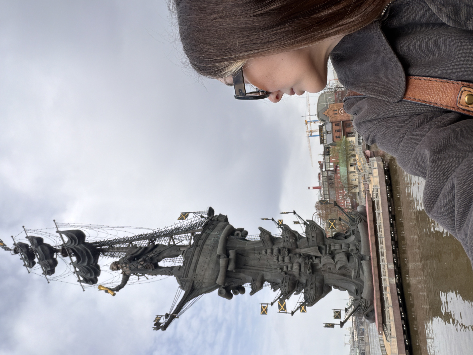 A person stands facing the Peter the Great statue in Moscow, Russia, looking down at the statue and the Moskva River it stands in. The statue depicts a ship with many flags. The backdrop includes buildings, some under construction. The sky is overcast. The person is wearing glasses and a jacket with a brown strap.
