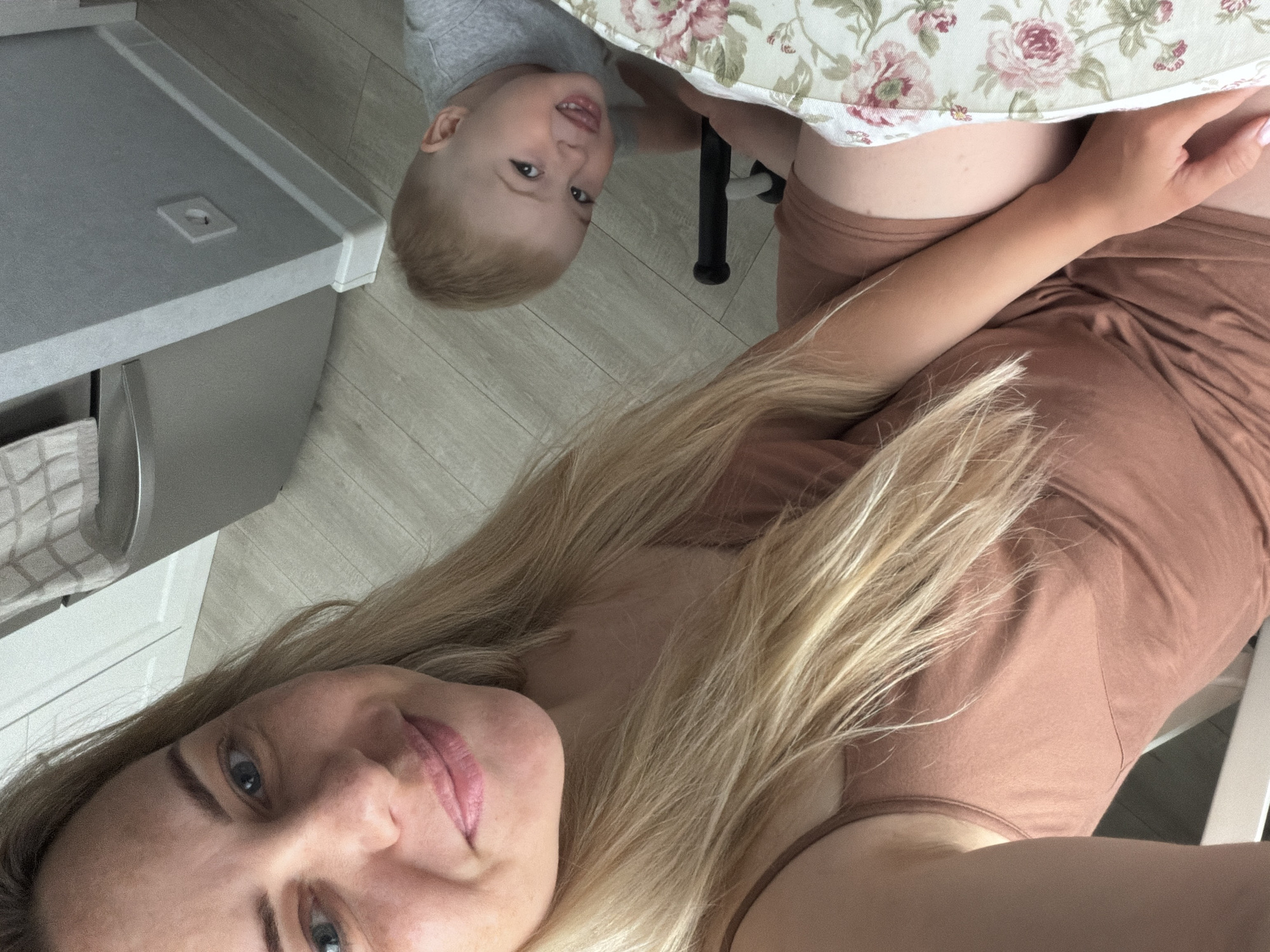 A woman with long blonde hair is taking a selfie. She is wearing a brown top. Next to her, under a floral patterned tablecloth, is a young child with blonde hair. The scene appears to be indoors, possibly a kitchen, with a counter and cabinets visible. The location in the city of Votkinsk, Russia, cannot be confirmed from the visual information provided.