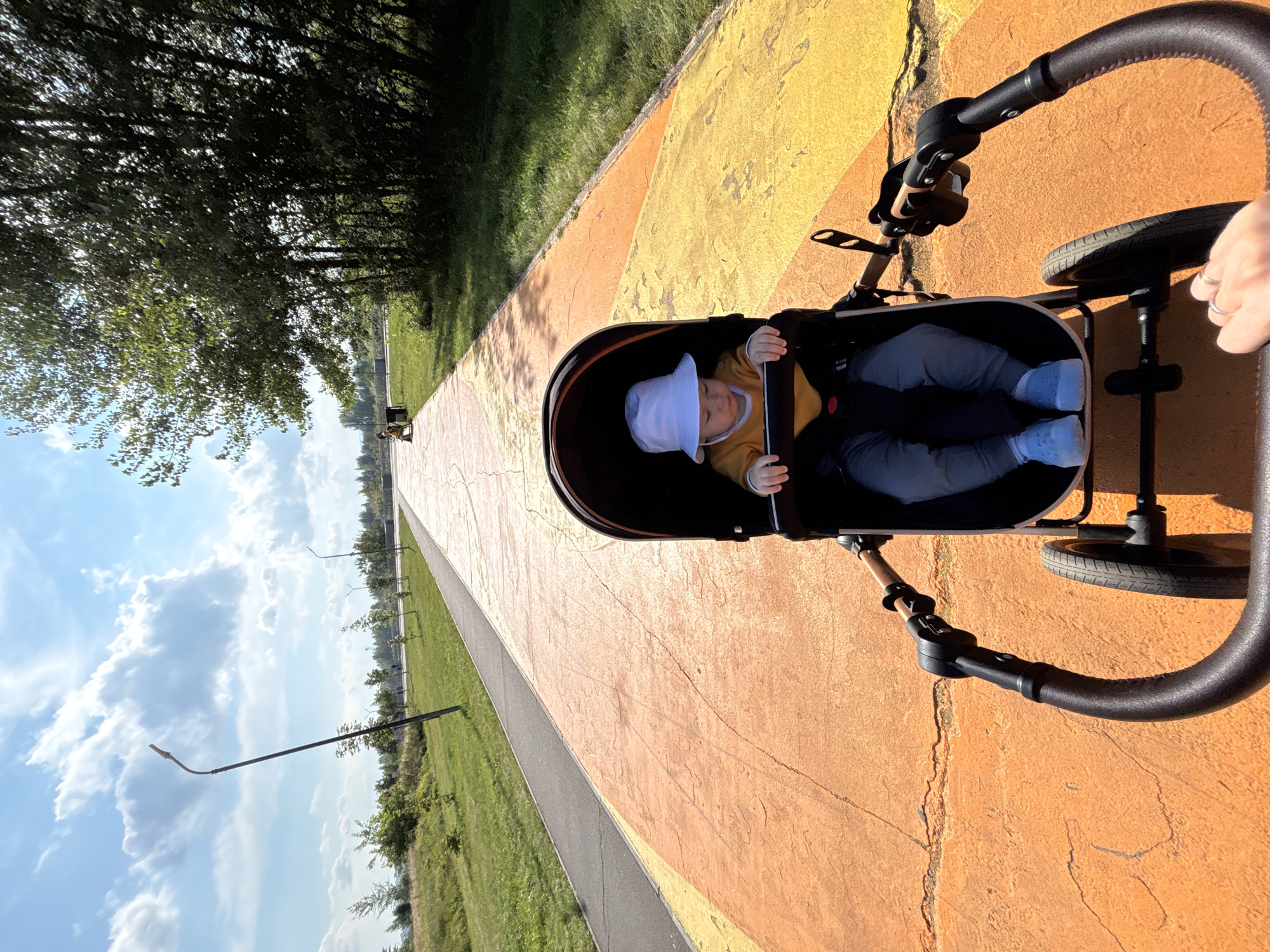 A baby wearing a yellow shirt and a white bucket hat sits in a stroller on a brightly colored orange and yellow path in a park. The path appears to be made of concrete with a textured surface. To the left of the path is a gray asphalt walkway and a grassy area with some trees. In the background, there are more trees and a blue sky with white clouds. A lamppost is visible on the left side of the frame. The stroller is being pushed by an unseen person, as indicated by a hand in the lower right corner holding the stroller handle. The scene suggests a pleasant day outdoors in a public park.