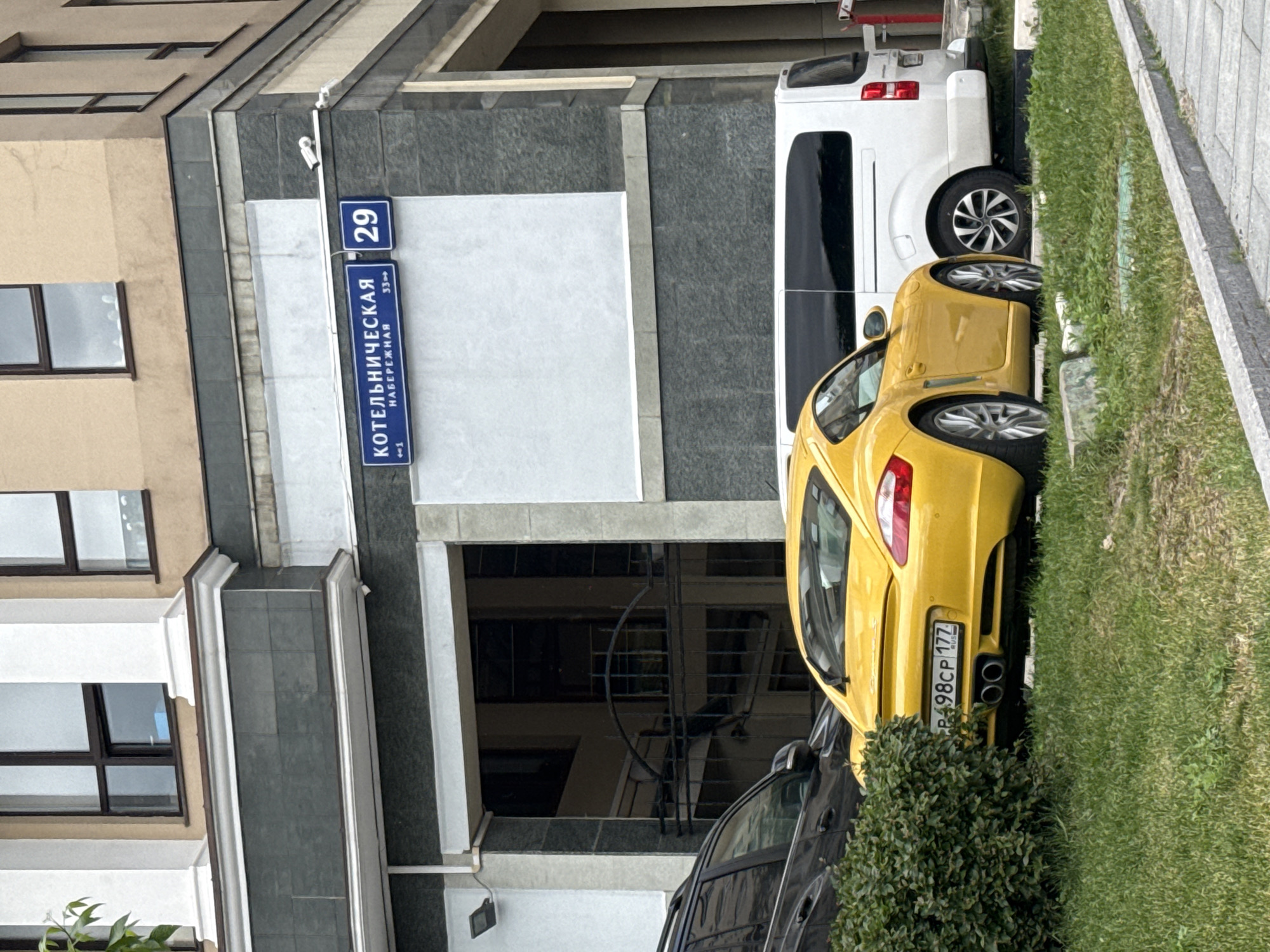 The image shows a street scene in Moscow, Russia, specifically at 29 Kotelnicheskaya Naberezhnaya. A yellow Porsche is parked on the right side of the image, near a patch of grass and a sidewalk. Next to the Porsche is a white van. A black car is also partially visible behind the Porsche. The building behind the cars displays the address sign.