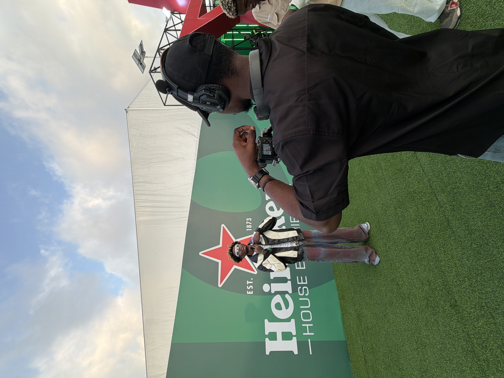 A man in a black shirt and baseball cap is holding a camera and filming another man standing in front of a large Heineken sign on green turf. The sign includes a red star and the text "EST. 1873 Heineken - HOUSE EXPERIENCE." The man in front of the sign is wearing sunglasses and a jacket. The sky is visible in the background. The image was captured at the Heineken House Experience in Lagos, Nigeria.