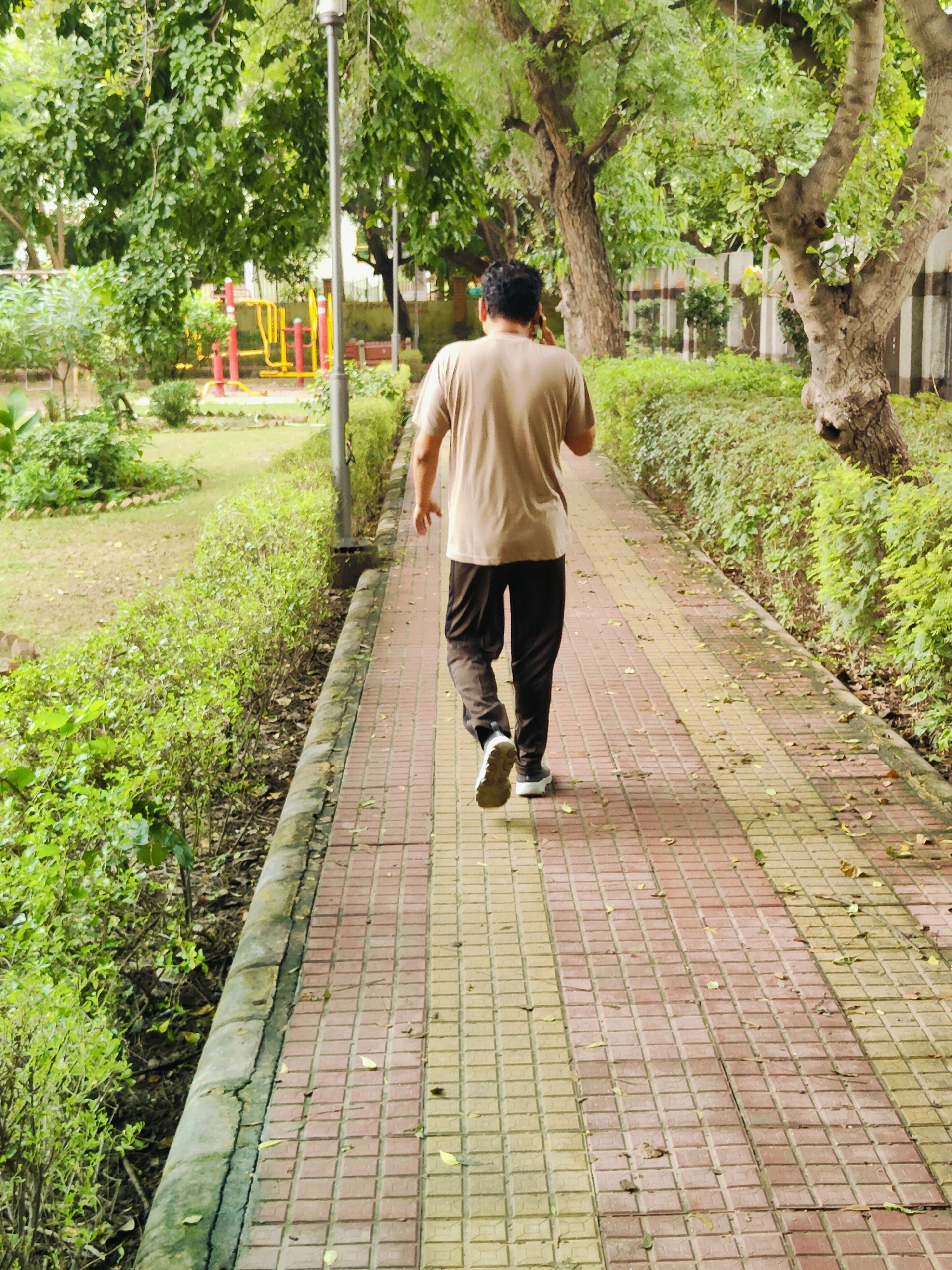 A man wearing a tan t-shirt and dark pants walks away from the camera on a paved pathway in a park. The pathway is made of alternating red and yellow square tiles. The man is wearing white athletic shoes. To the left of the pathway is a grassy area with some low green bushes, and further in the background are trees and a red and yellow playground structure. To the right of the pathway is a dense hedge of green bushes, and beyond that, a tree trunk and a building. The scene appears to be outdoors in a public park, likely in Gurgaon, India, given the lush greenery and presence of park amenities. The lighting suggests it is daytime, possibly overcast, as there are soft shadows.