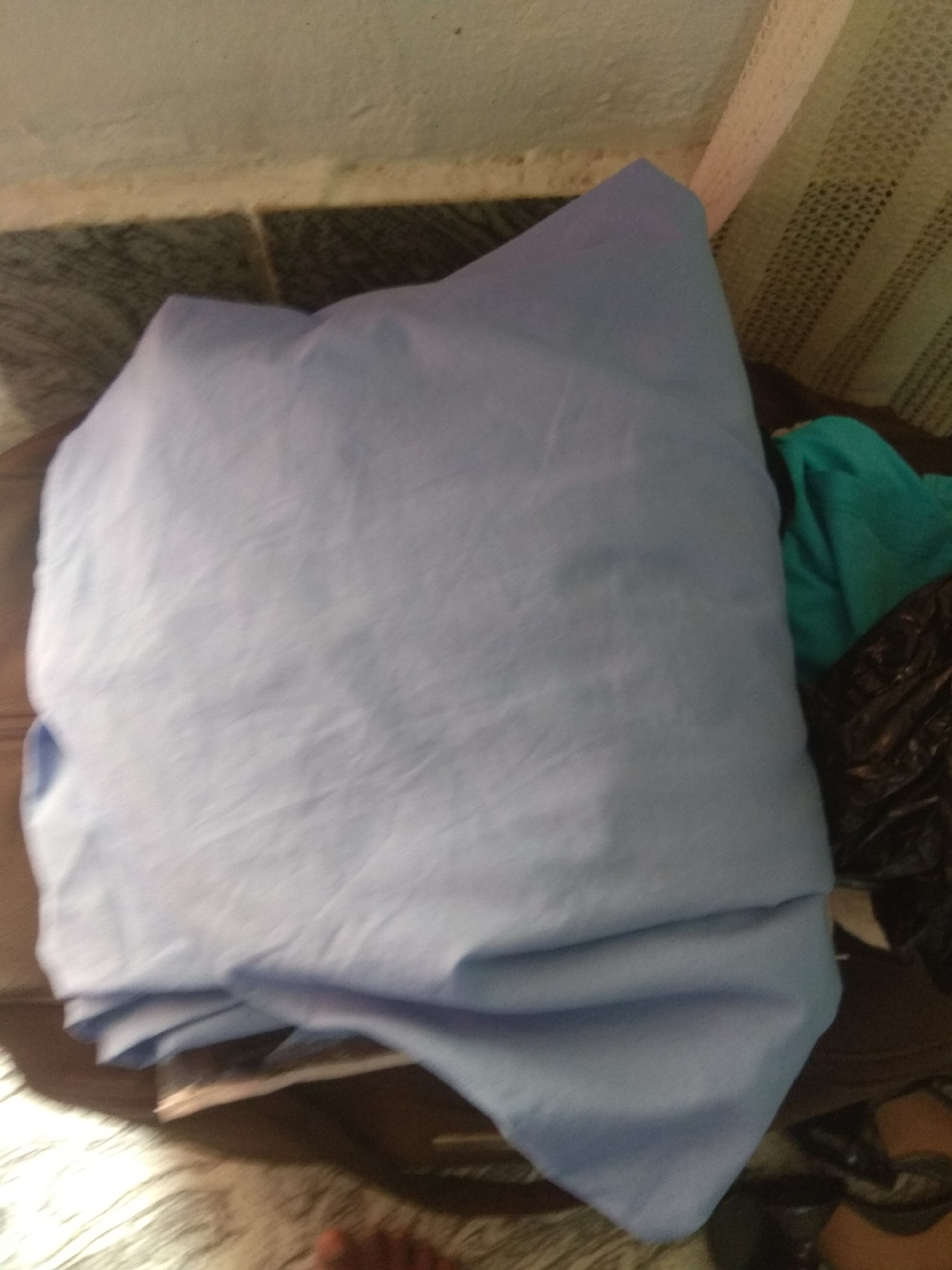 The media file shows a pile of light blue fabric, likely sheets, in a domestic setting in Jalingo, Nigeria. The fabric is draped and folded, creating a soft, textured surface with visible wrinkles and creases. To the right of the fabric, a teal-colored cloth and a dark, crinkled plastic bag are partially visible, suggesting other items are present in the vicinity. The background shows a tiled floor and what appears to be a mosquito net, a common item in Nigerian homes. The lighting is dim, indicating an indoor scene, possibly during the evening or in a poorly lit room. No people are clearly depicted, although a foot is partially visible at the bottom of the frame, suggesting a person is nearby. The overall impression is of laundry or stored textiles in a casual, everyday environment. No text is visible in the image.
