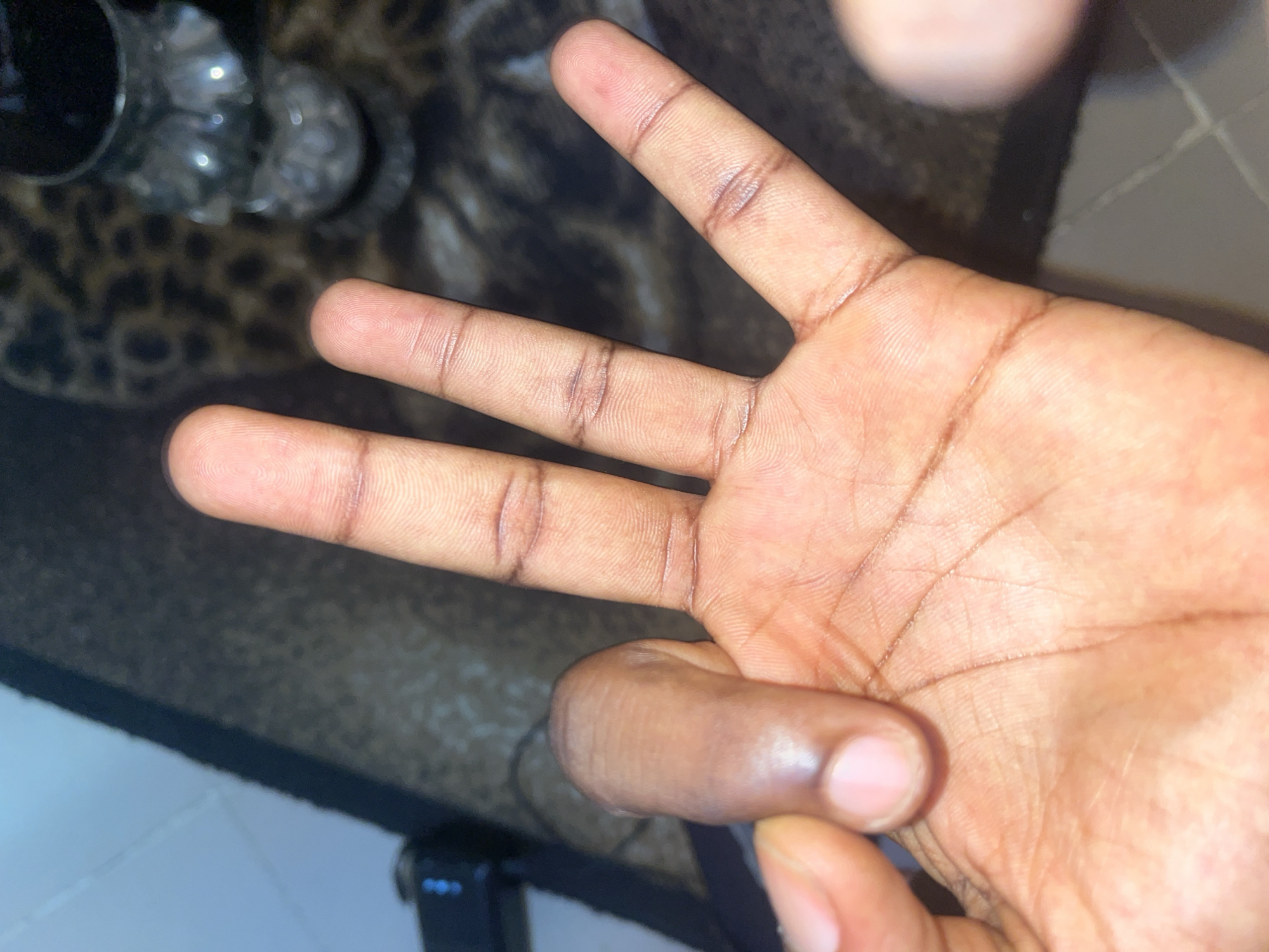 A close-up, overhead shot shows a person's left hand, palm facing upwards. Three fingers are extended, and the thumb is bent inward, obscuring part of the palm. The skin appears dark and shows creases and faint lines. In the background, out of focus, is a patterned rug and a glass with ice cubes. The lighting suggests an indoor setting. The image's metadata indicates it was taken in Jalingo, Nigeria.