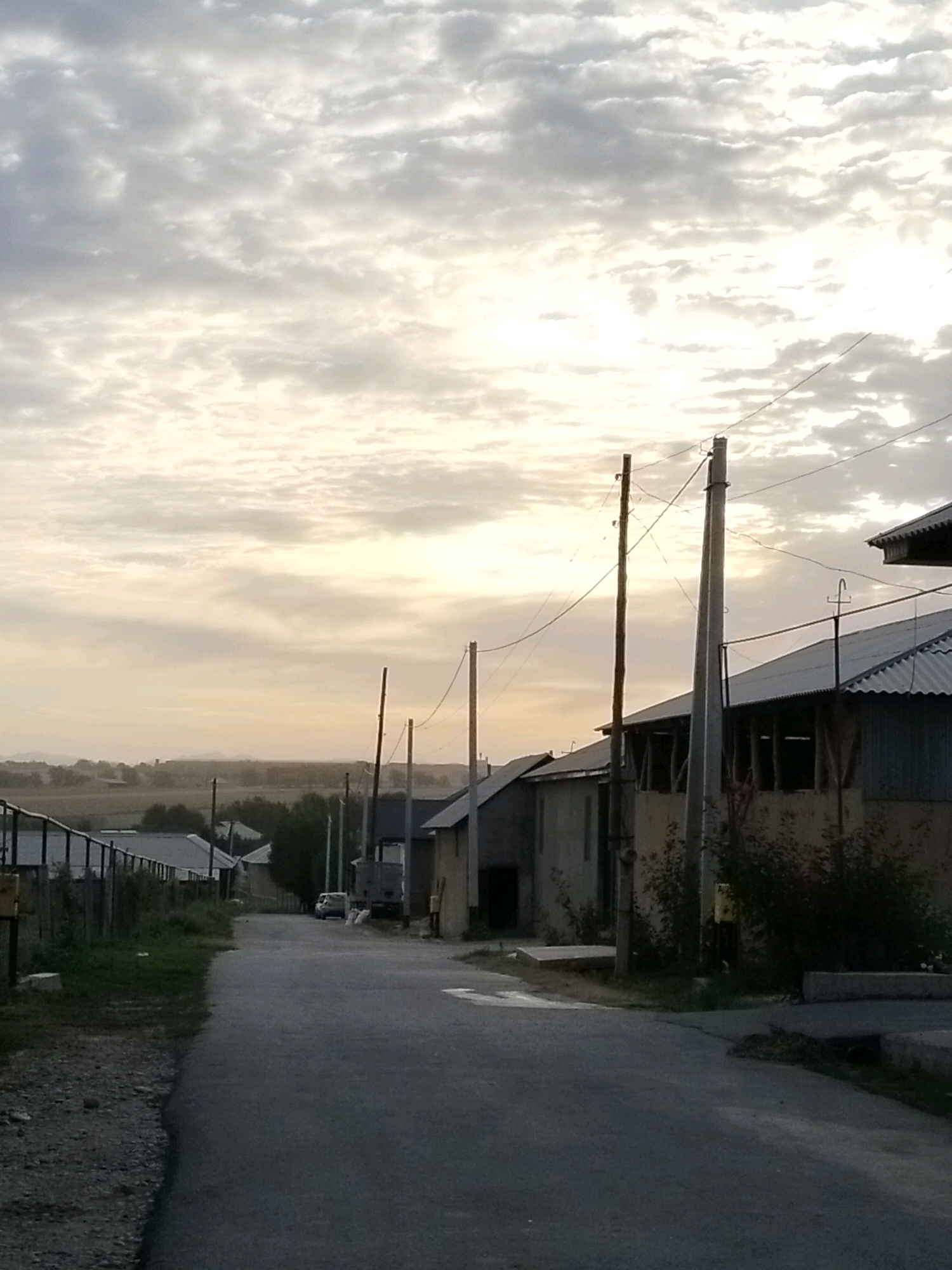 The image depicts a street scene in the city of Kölkent, Kazakhstan. The view is from a low angle, looking down a paved road lined with houses and utility poles. The sky is overcast with a light gradient from bright near the horizon to darker clouds above. On the left side of the road, there is a fence and some vegetation. Several houses with corrugated metal roofs are visible along the right side of the street, some with open-air structures attached. Utility poles with wires are prominent throughout the scene. A white vehicle is parked further down the road on the right. The overall impression is of a rural or semi-urban residential area under a cloudy sky.